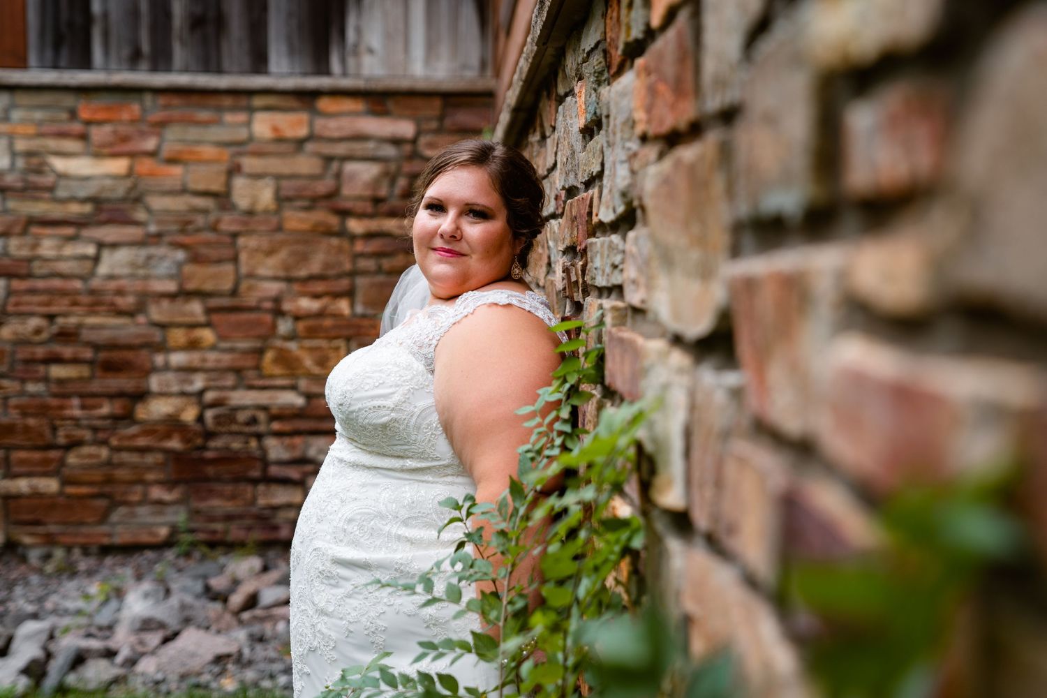 Bride in lace wedding gown poses elegantly beside rustic brick wall with green ivy accents.