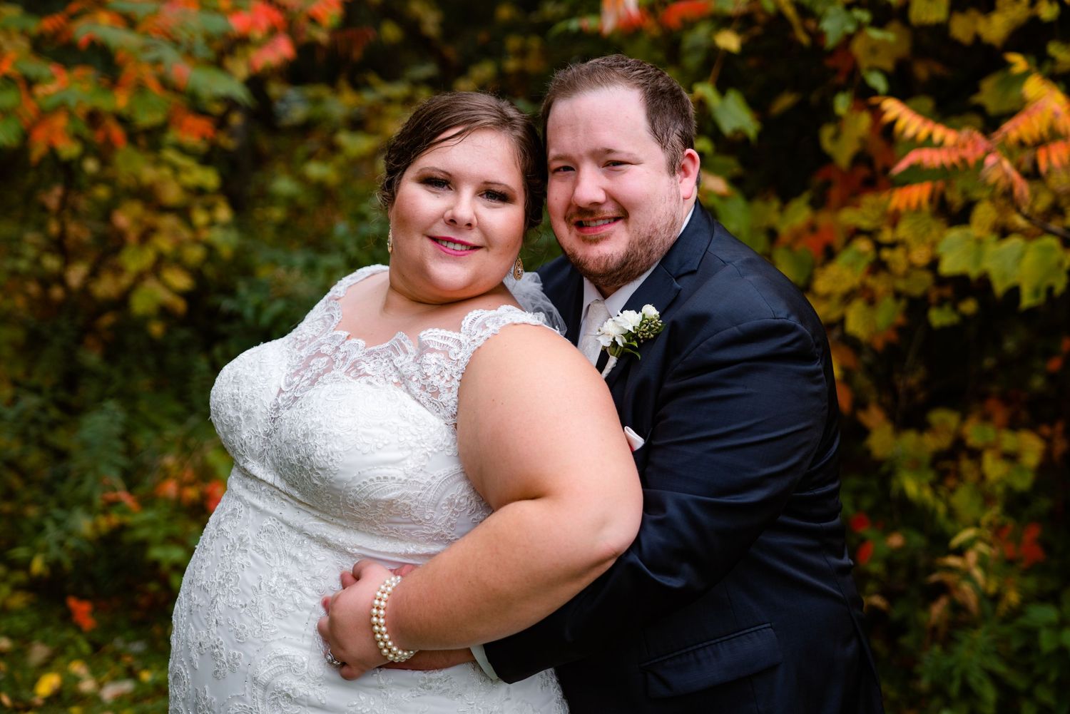 Happy bride and groom embracing in front of vibrant autumn foliage on their wedding day.