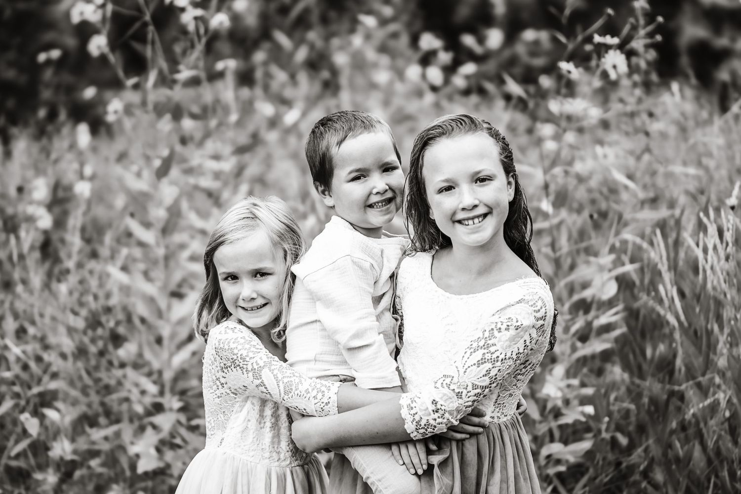 Black and white photo of three happy siblings hugging in a wildflower meadow outdoors.