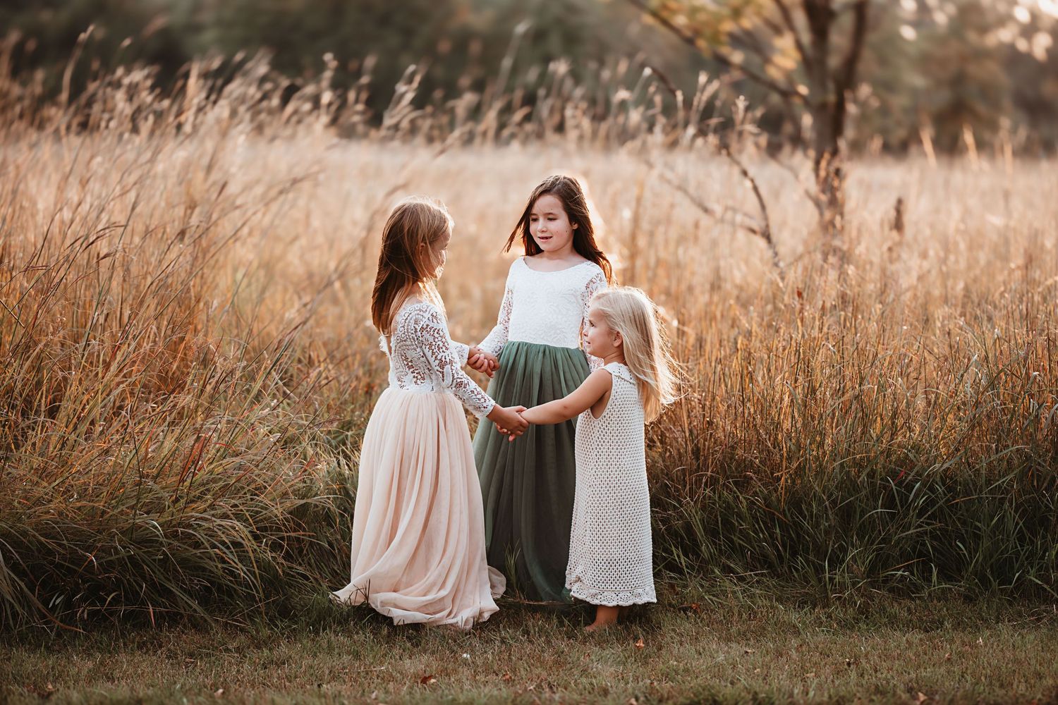 Three girls in flowing dresses hold hands in a golden field at sunset.