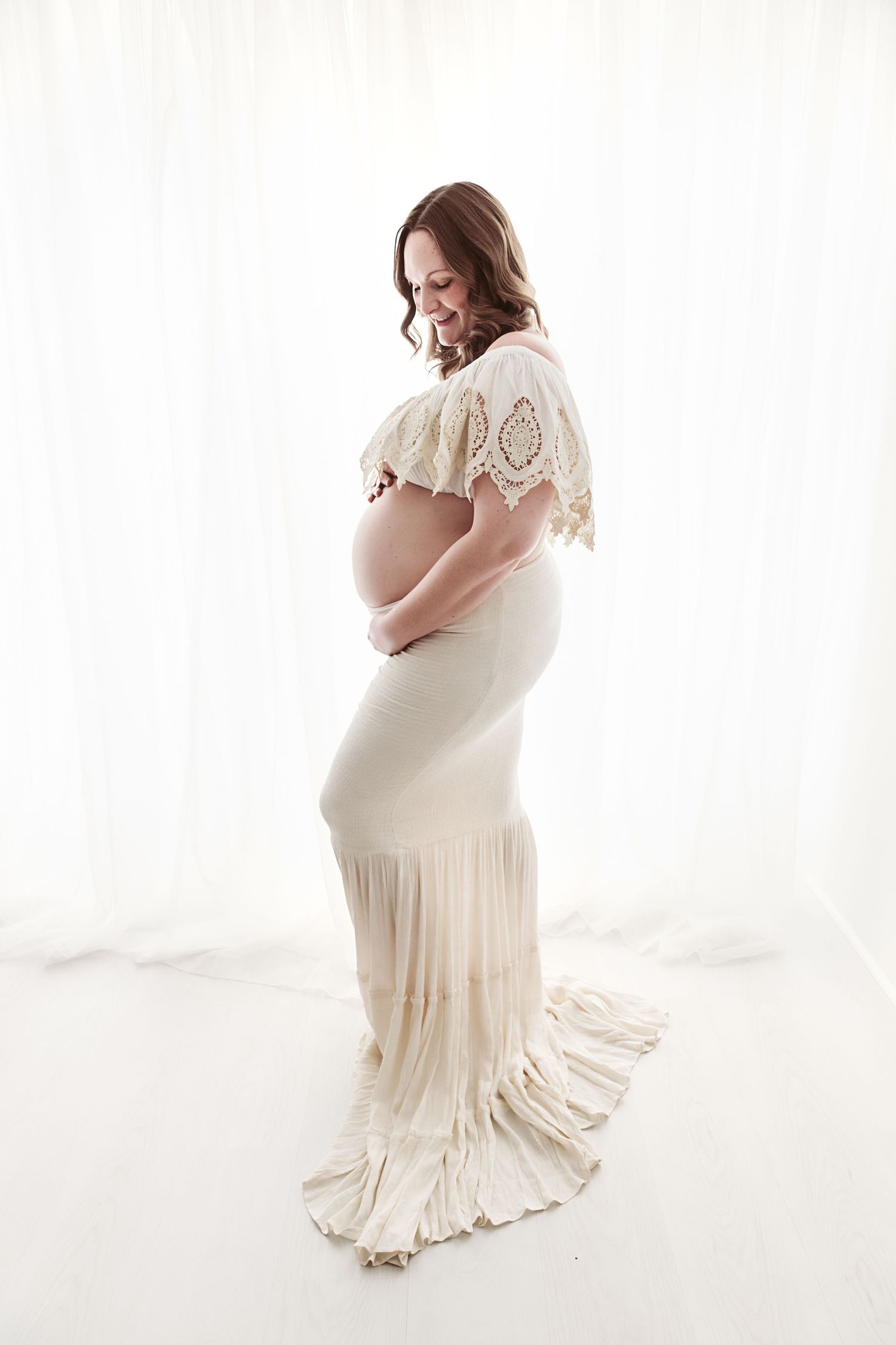 Pregnant woman in elegant cream lace gown smiling down at her baby bump in white studio.
