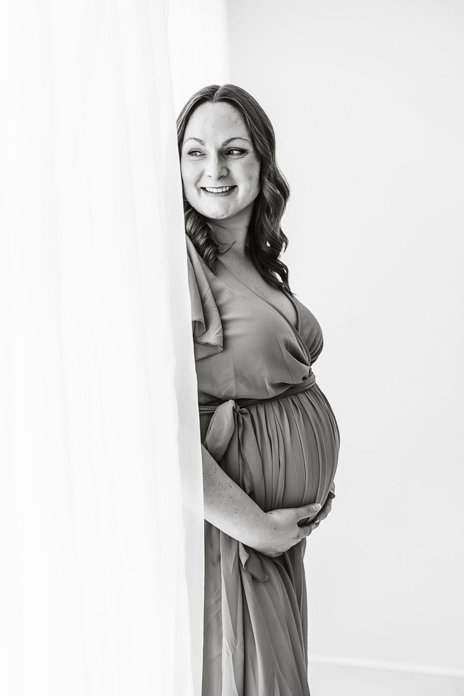 Smiling pregnant woman in flowing dress peeking around white curtain, black and white maternity portrait.