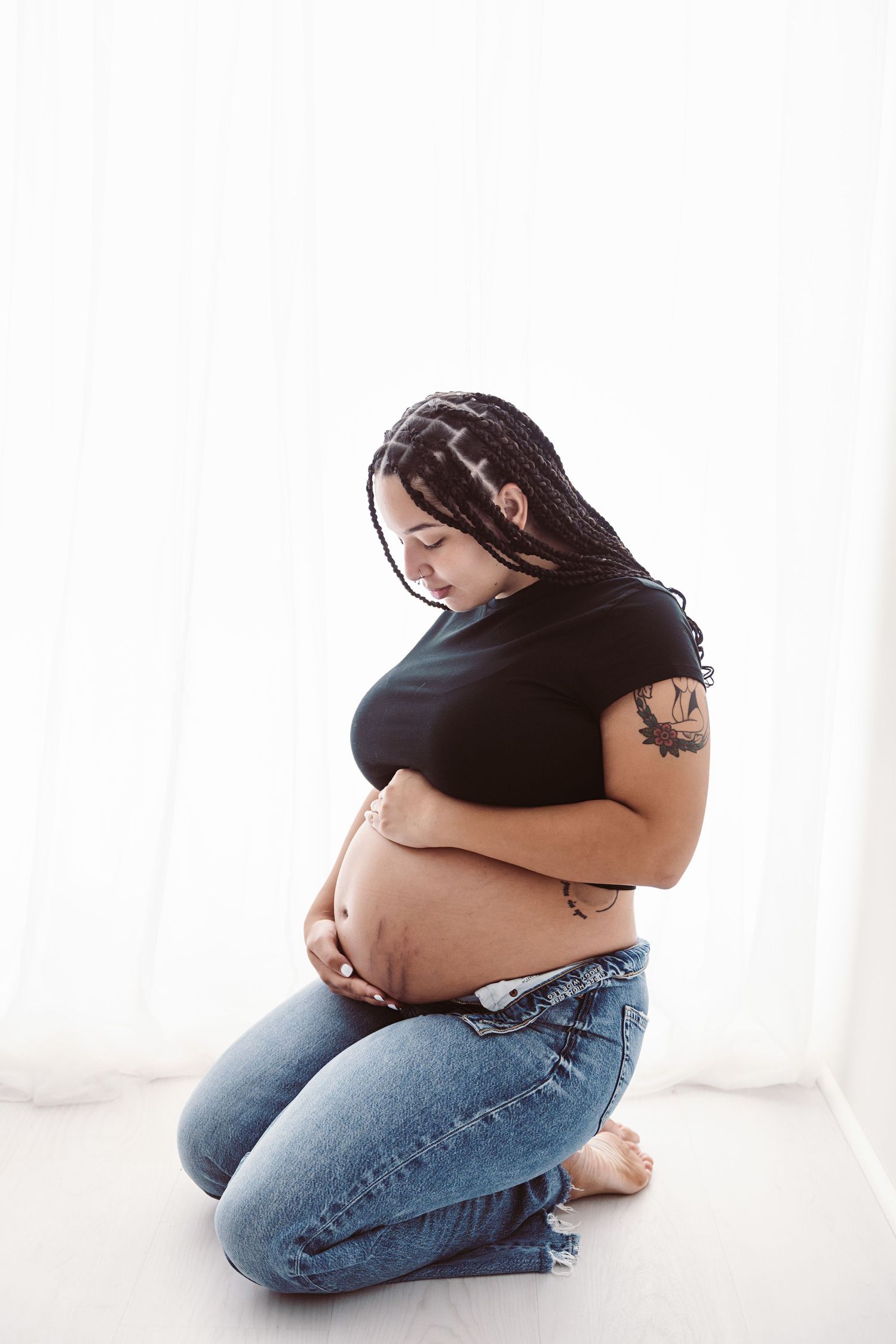 Pregnant woman with braids kneeling on floor cradling her bare baby bump in black top and jeans.
