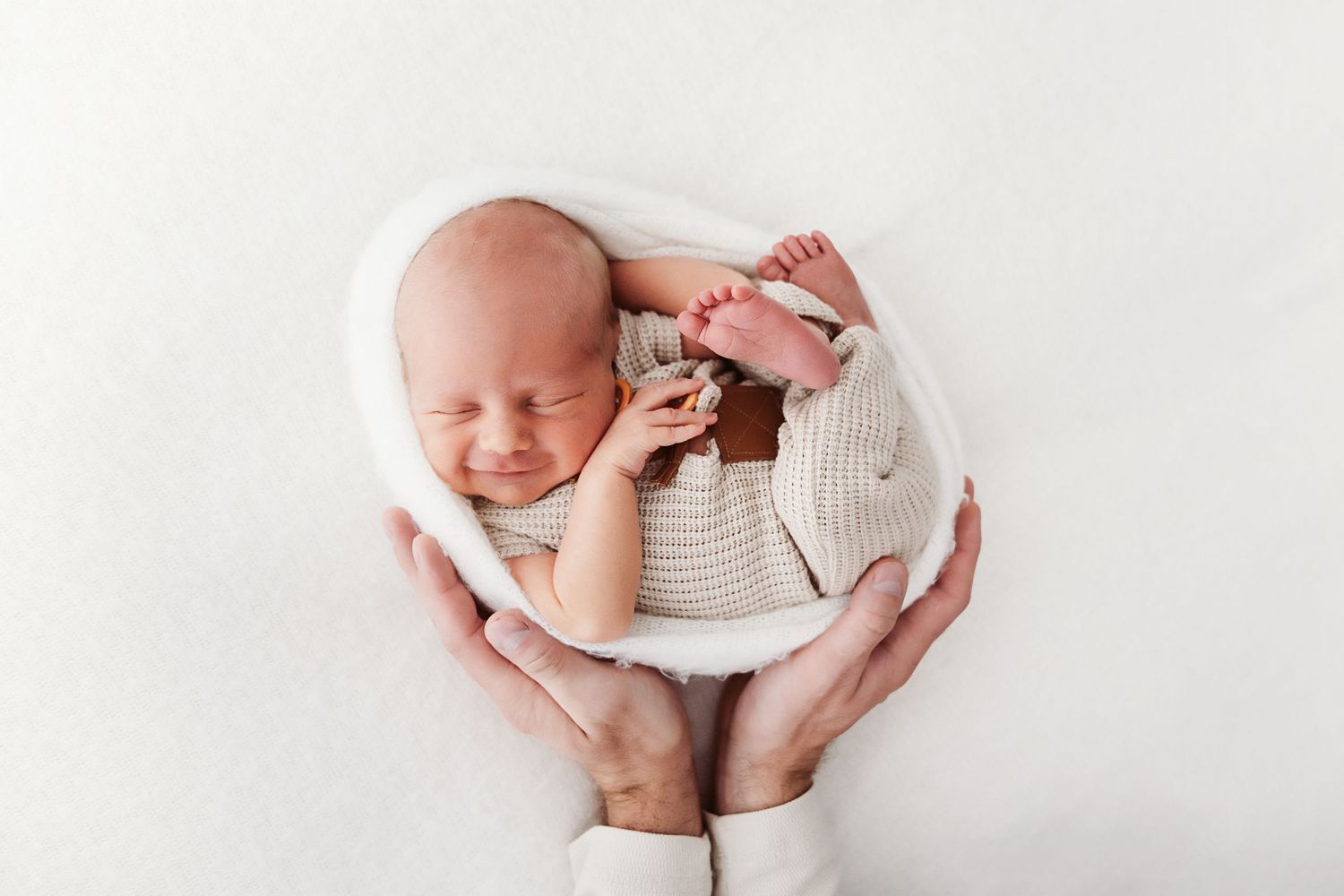 Smiling newborn in knit outfit cradled gently in parents hands on white blanket.