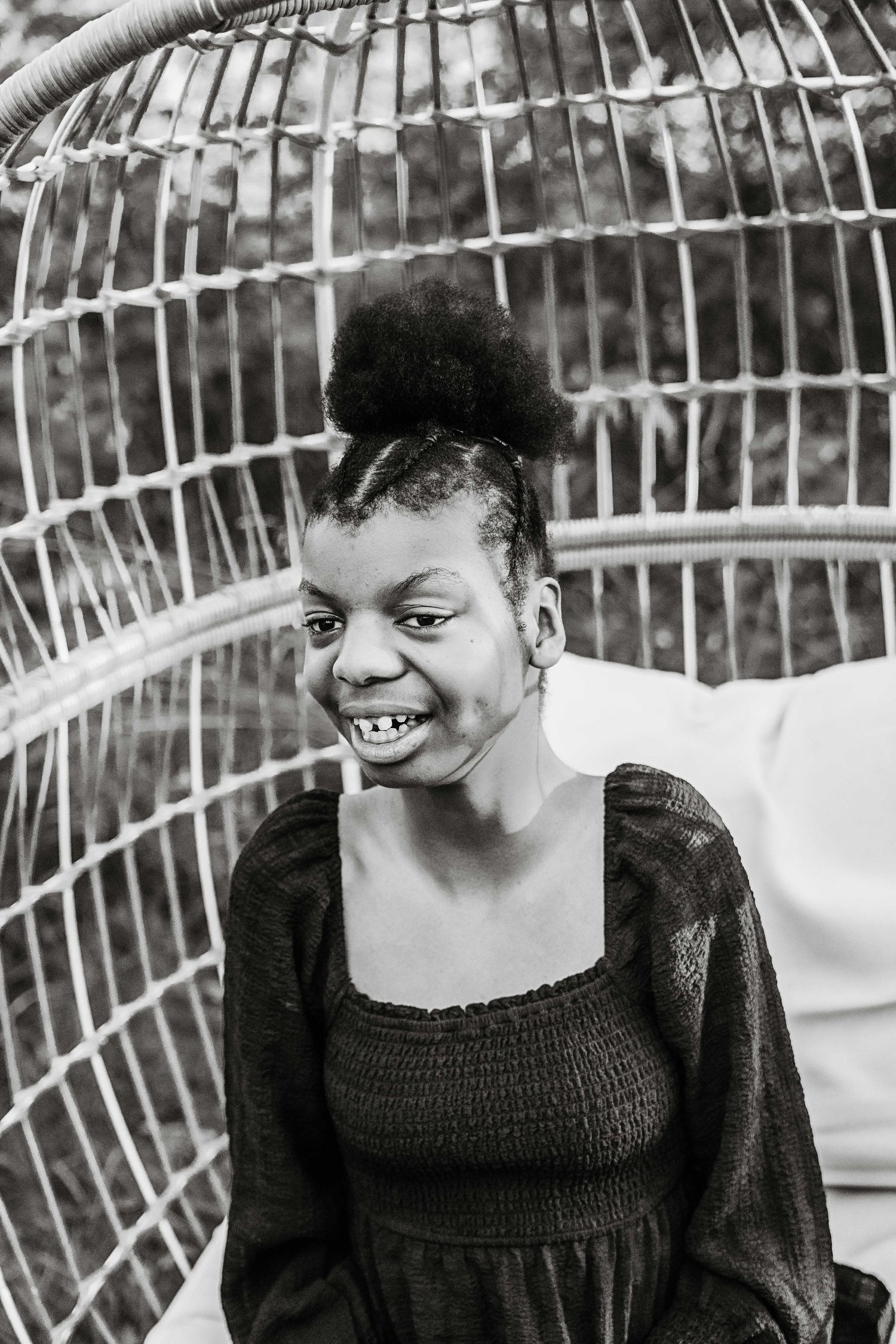 Black and white portrait of a young girl smiling in a wicker egg chair outdoors.