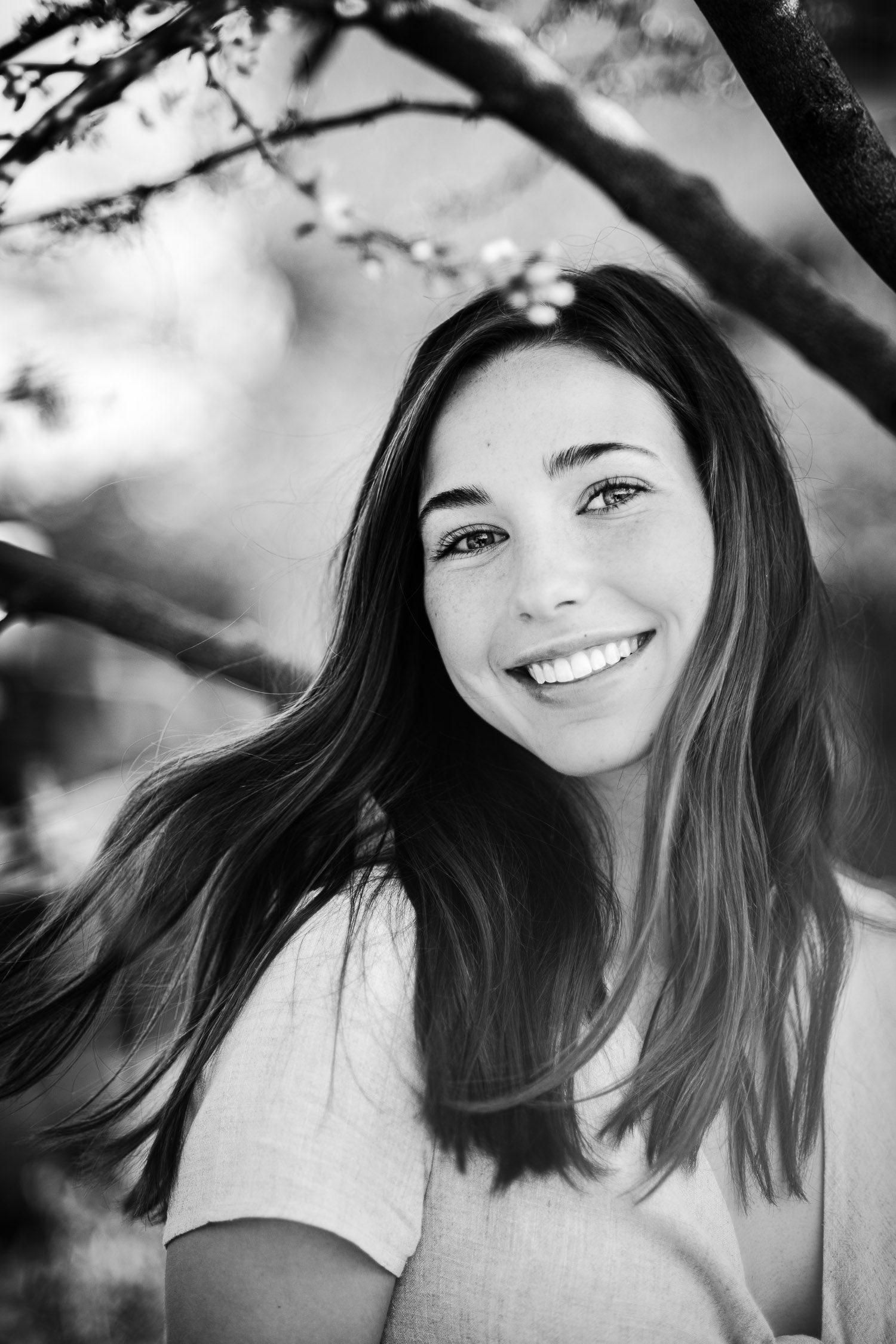 Black and white portrait of a smiling young woman with long hair outdoors under flowering tree branches.