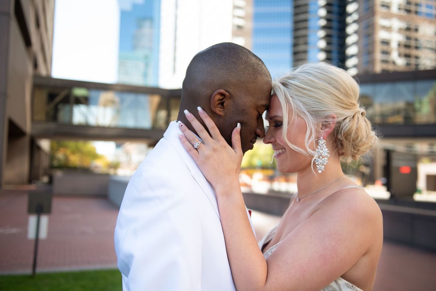 Interracial wedding couple sharing an intimate forehead touch moment outdoors with city buildings in background.