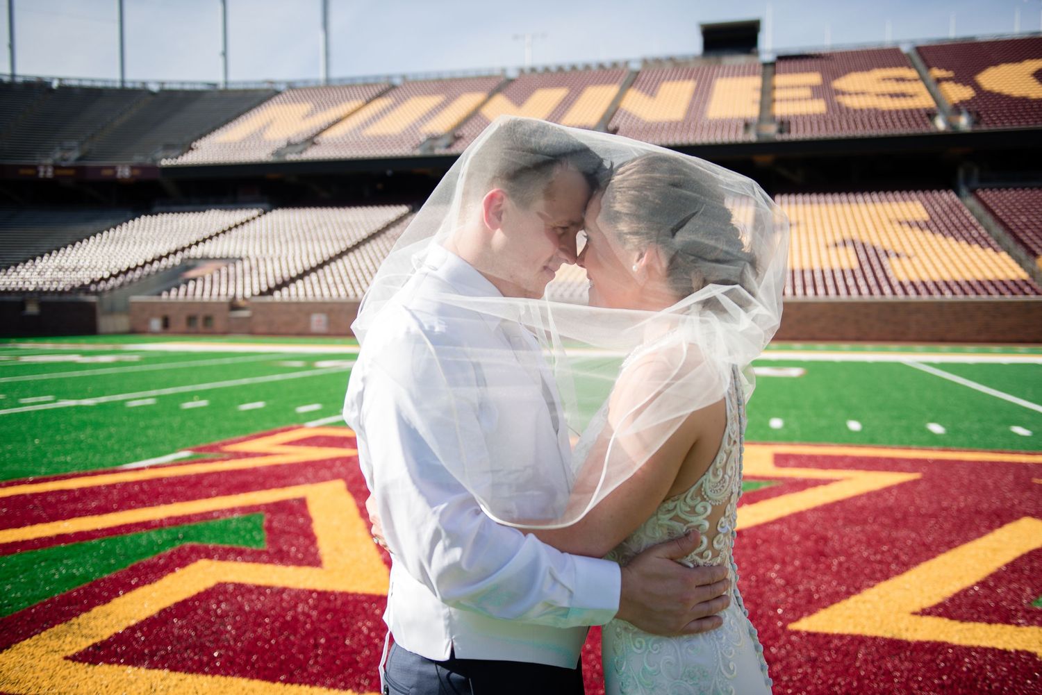 Bride and groom sharing an intimate kiss under a veil on a college football field.