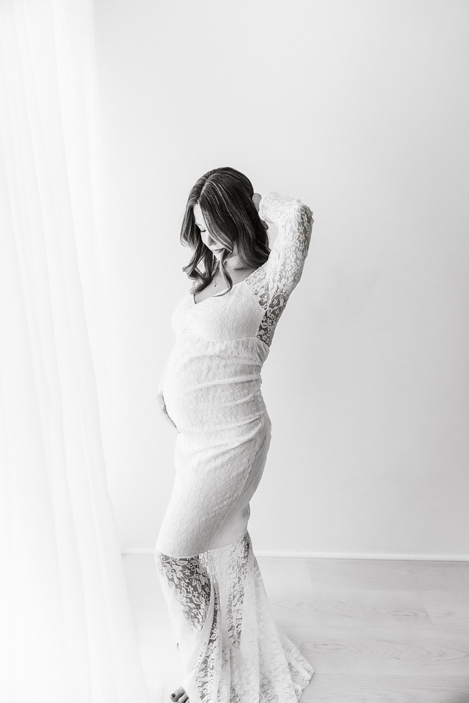 Black and white portrait of pregnant woman in white lace gown posing gracefully by a window.