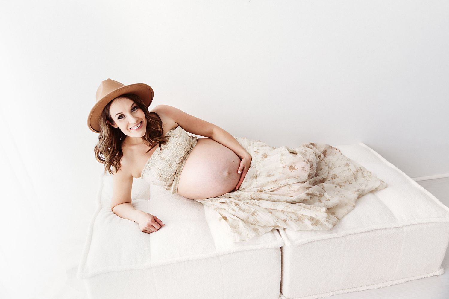Smiling pregnant woman in floral dress and tan hat lying on white couch in bright studio setting.