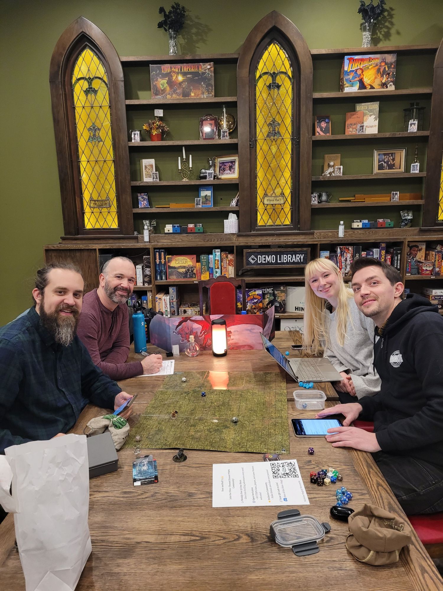 four individuals smiling surrounding ttrpg gaming table with stained glass shelves in the background