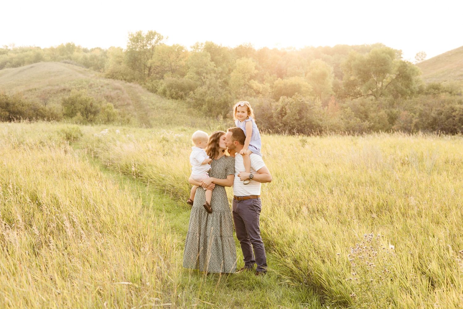 A family stands together in a sunlit golden meadow during sunset with rolling hills in the background.