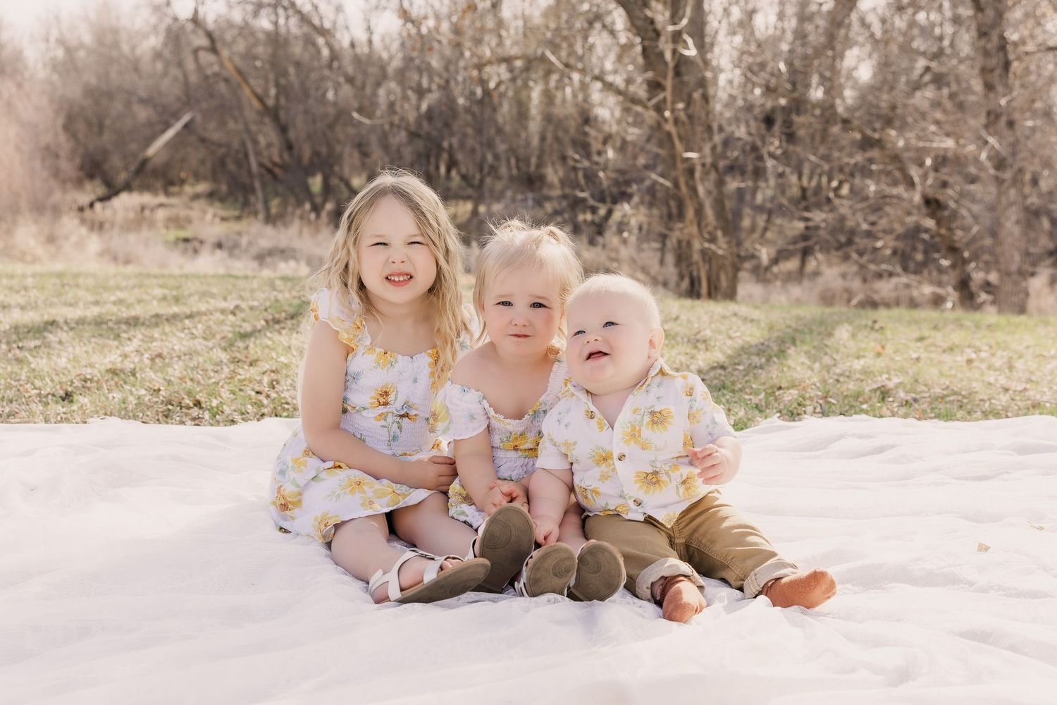 Three young children sitting together on a white blanket outdoors wearing floral and neutral colored outfits.