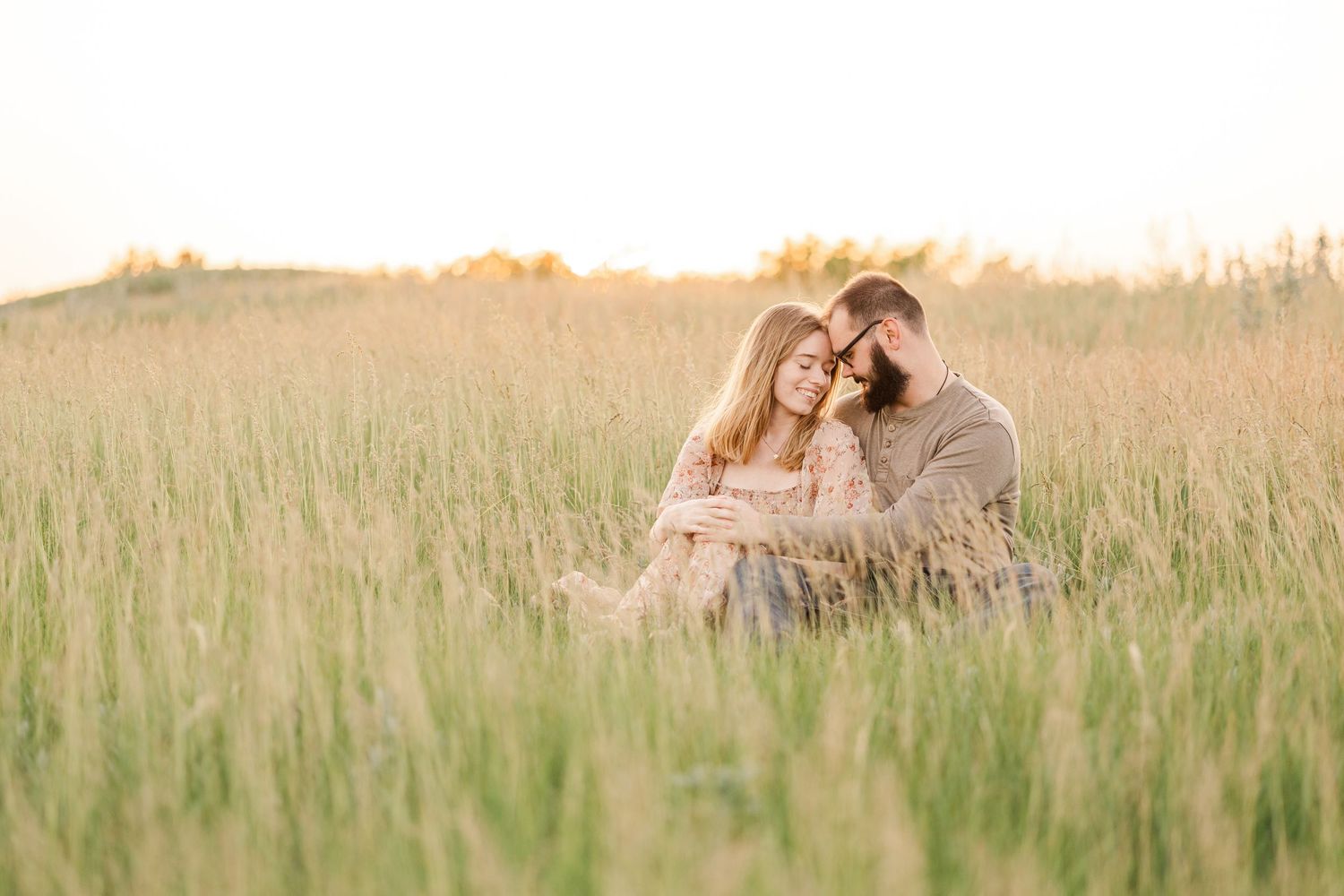 A couple sits intimately together in a golden wheat field during sunset, surrounded by tall grass swaying in the breeze.