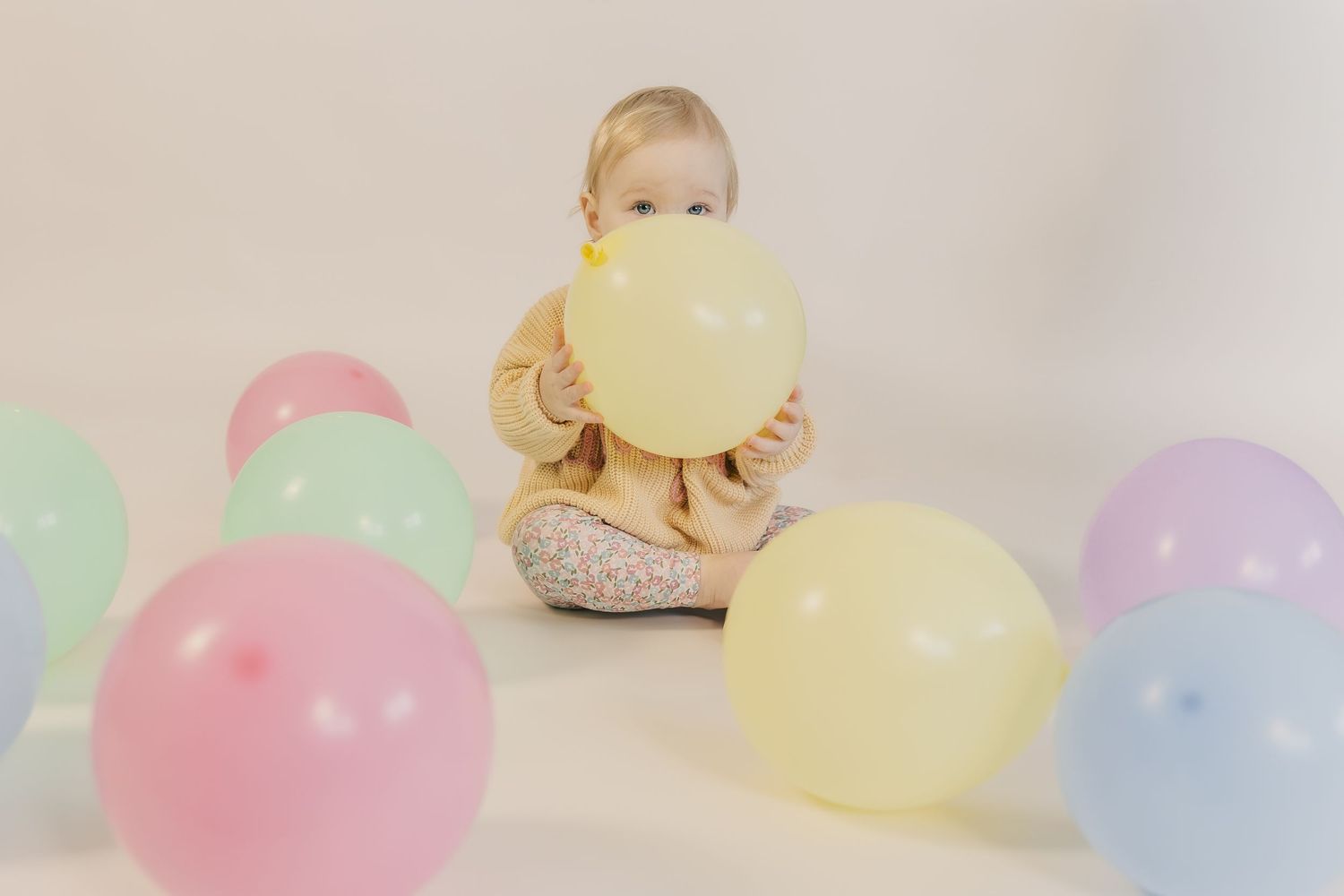 A baby sits on a white background surrounded by colorful pastel balloons, holding a yellow balloon.