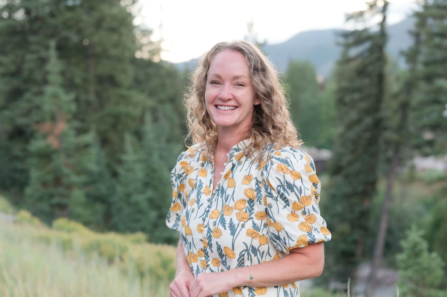 Smiling woman with curly blonde hair wearing a floral dress outdoors with pine trees and mountains.