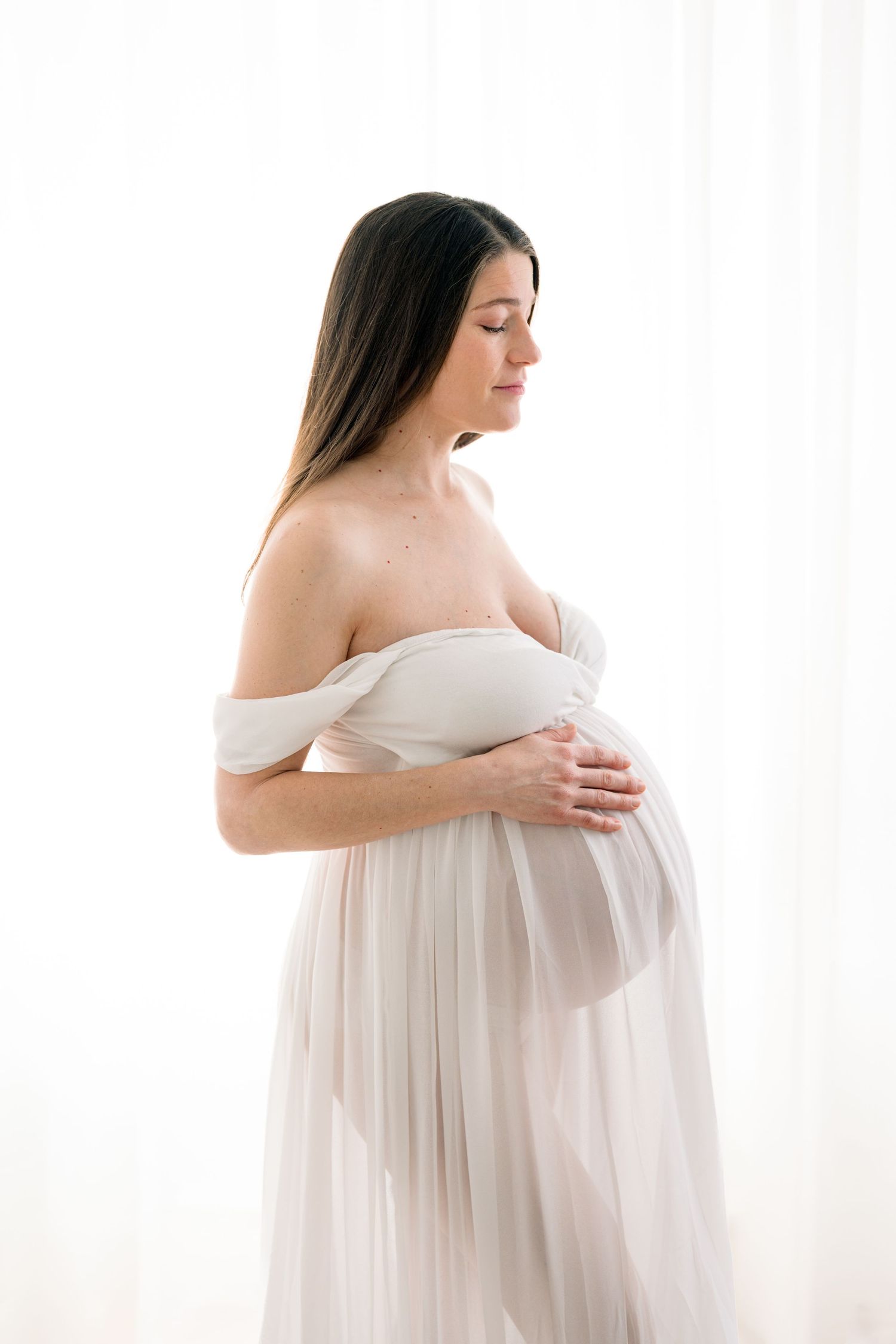Future maman dans une robe blanche et vaporeuse de la garde-robe du studio qui laisse voir sa silhouette de madone dans la lumière filtrée