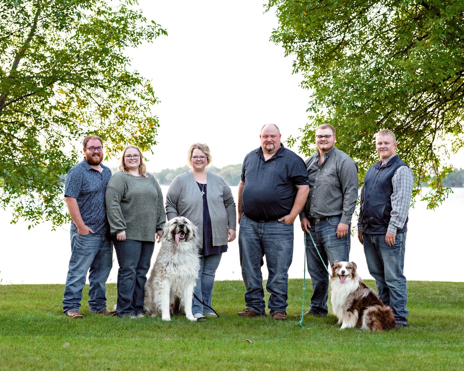 Family group of six adults poses outdoors with two dogs near a scenic lakeside willow tree.