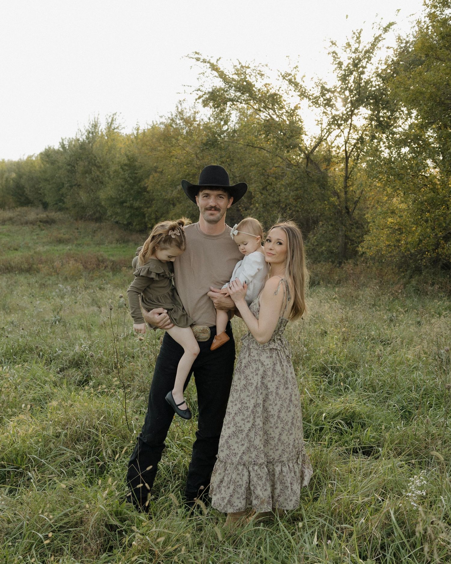 A family of four poses in a green meadow, man in cowboy hat holding toddler, woman in floral dress holding baby.