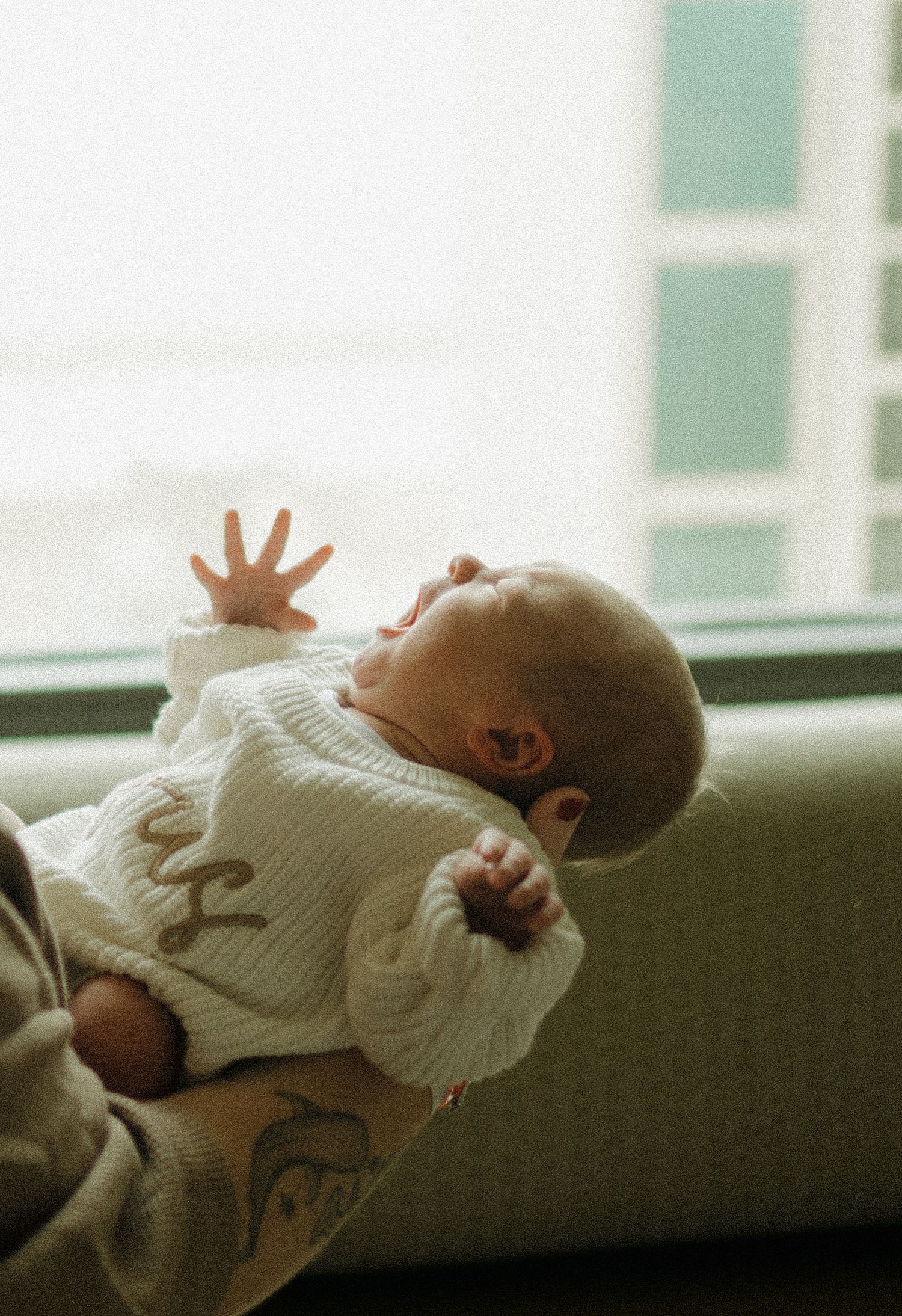 A newborn baby in a white knit sweater being held near a window, reaching one hand upward.