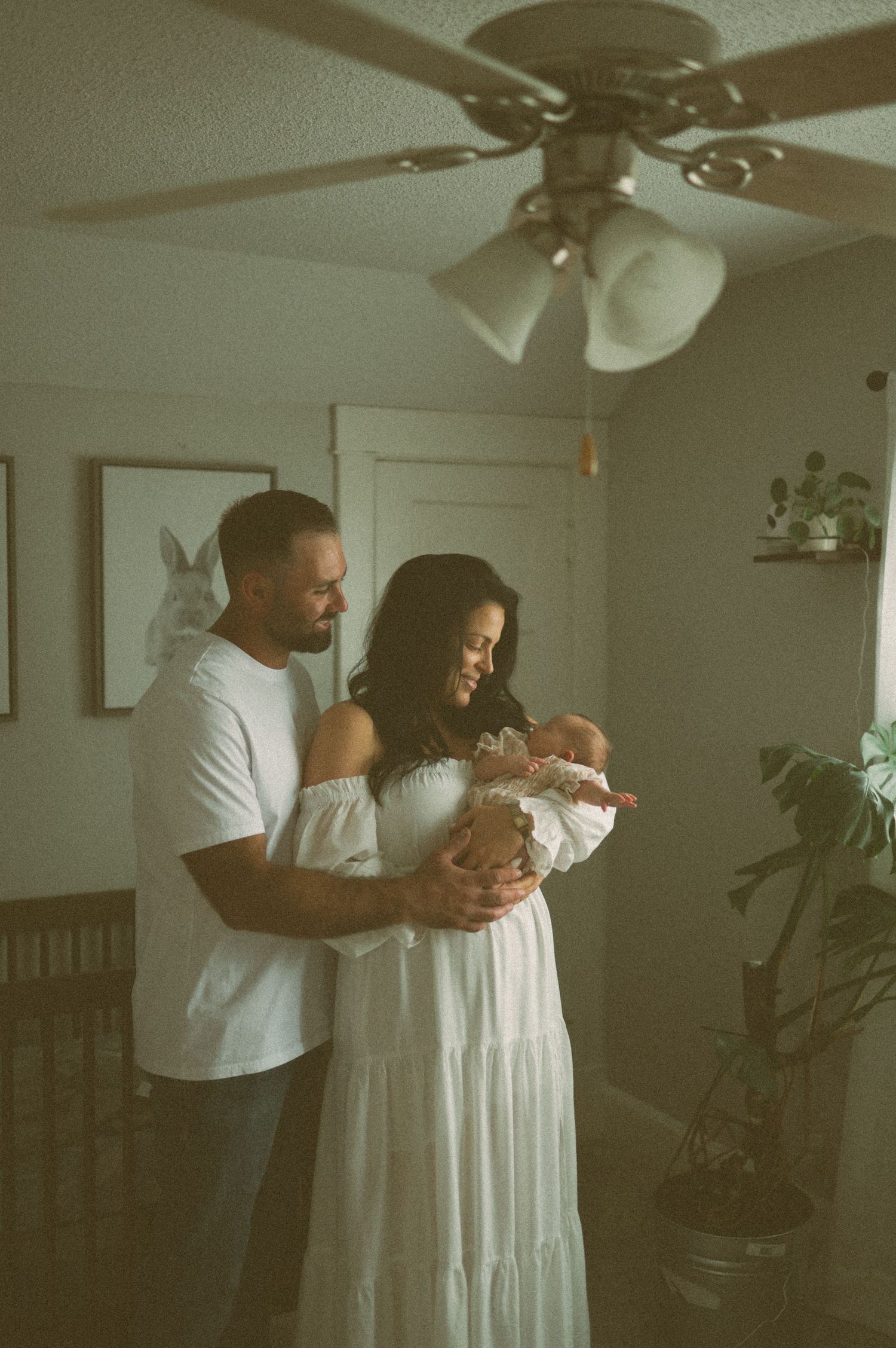 A couple in a softly lit room tenderly holds their newborn baby, the mother in a white off-shoulder dress.
