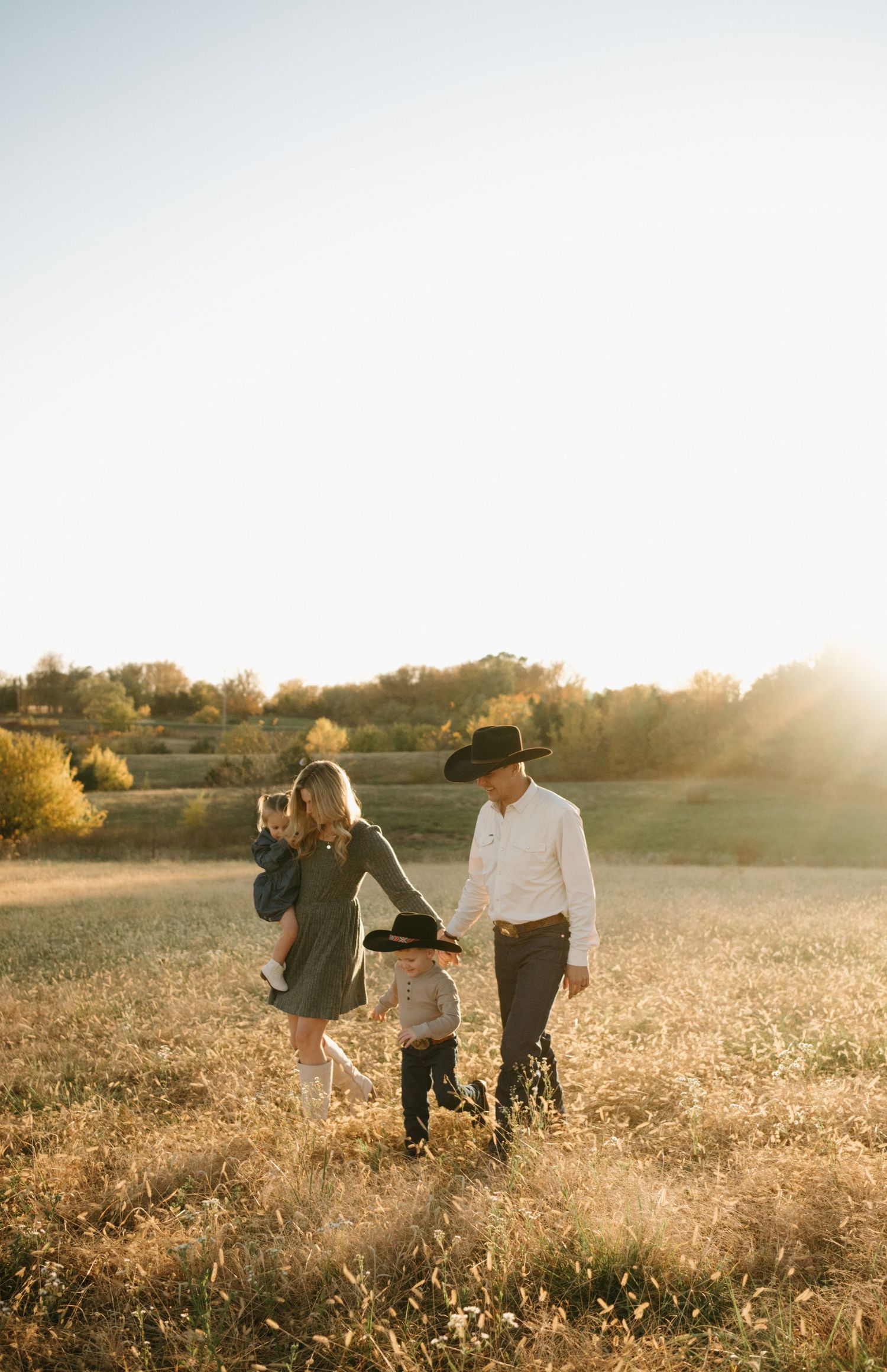 Family walking in golden field at sunset, parents in cowboy hats with two young children, warm autumn countryside backdrop.