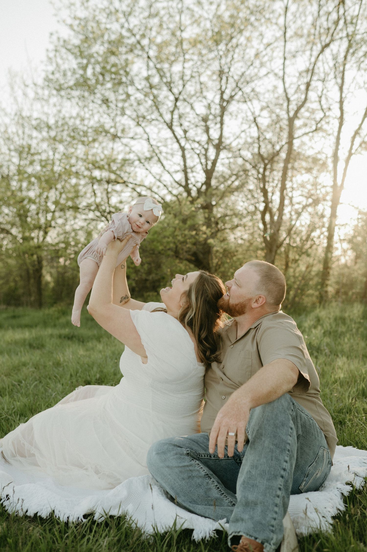 Young family sits on blanket in sunny green field, mom lifts smiling baby while dad kisses her cheek.