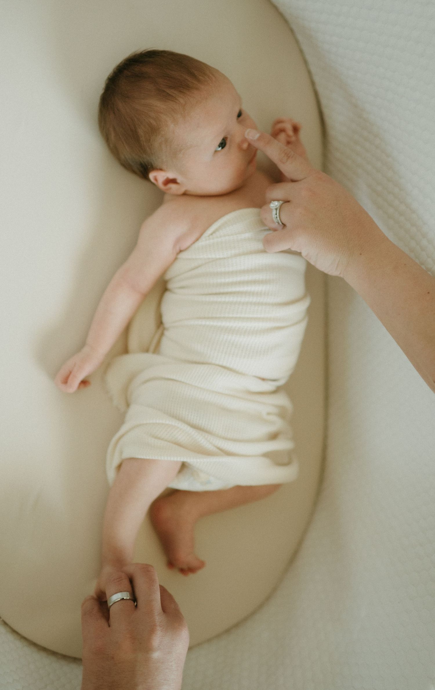 Newborn baby wrapped in cream knit fabric lying in a bassinet while a parent gently touches their cheek.