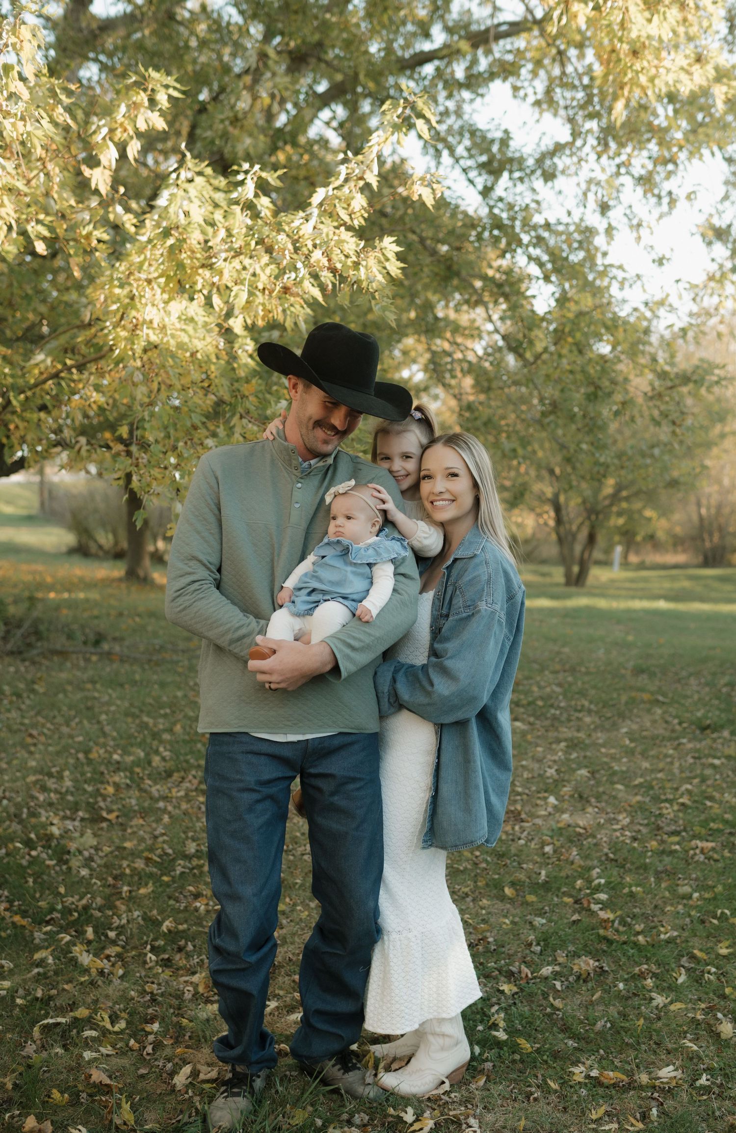 Smiling family of four poses under autumn tree, dad in cowboy hat holds baby and toddler daughter.