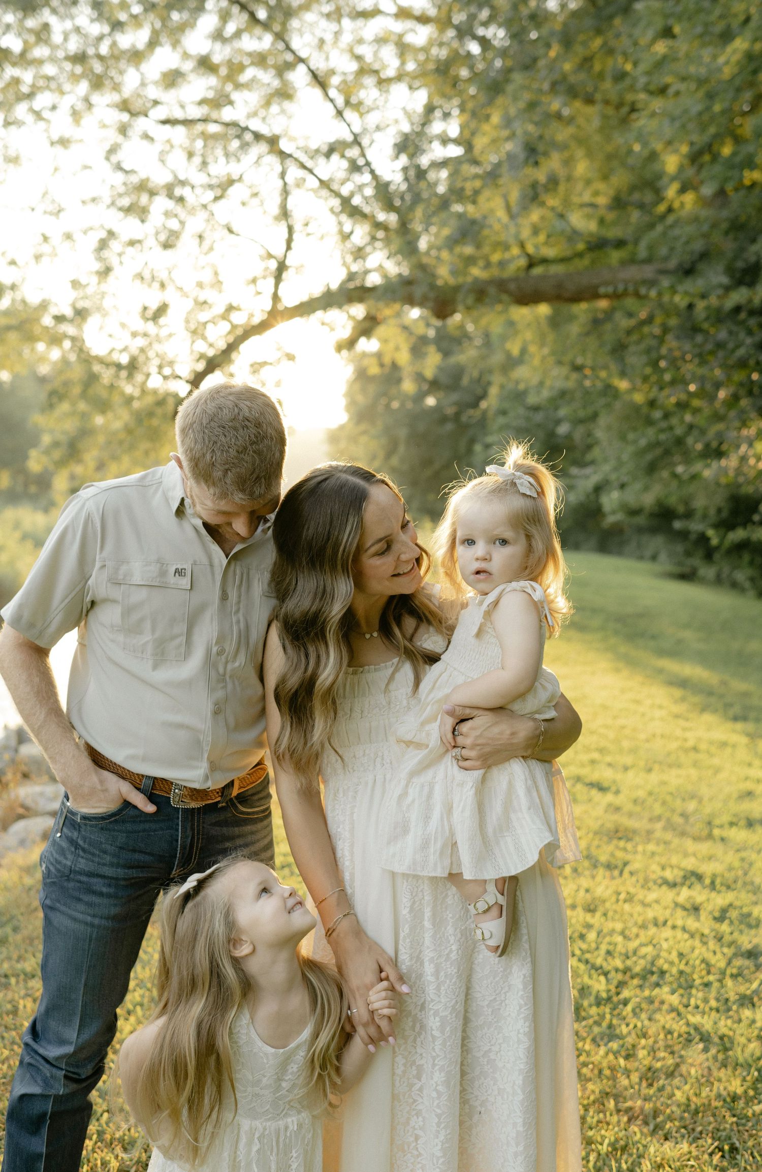 Family of four poses outdoors in golden hour light, parents smiling at toddler while young girl looks up adoringly.