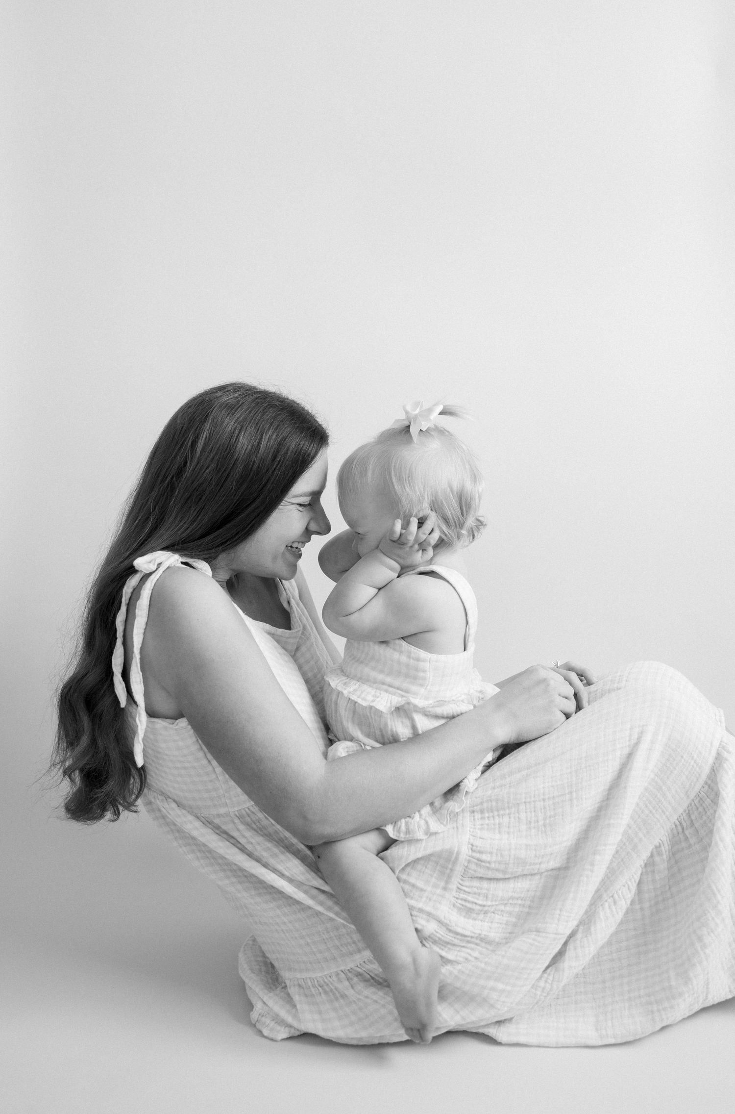 Black and white portrait of brunette mother in sundress sitting and cuddling her baby girl tenderly.