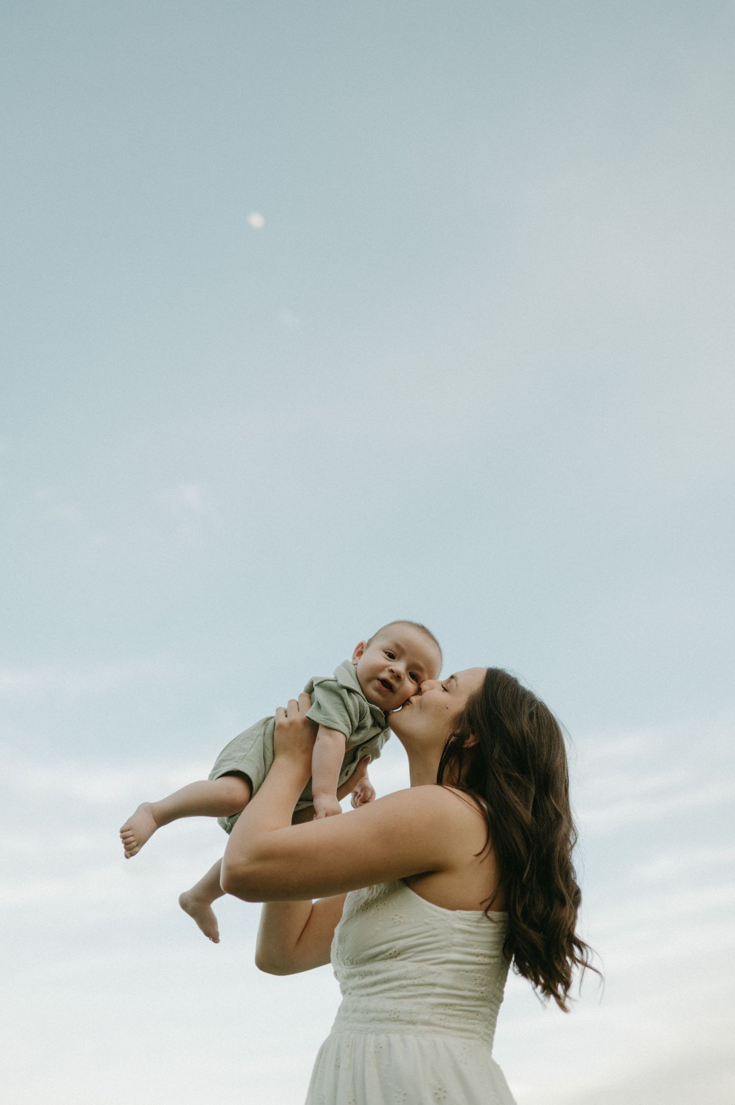 A joyful mother in a white dress lifts her laughing baby toward a bright blue sky in an outdoor setting.