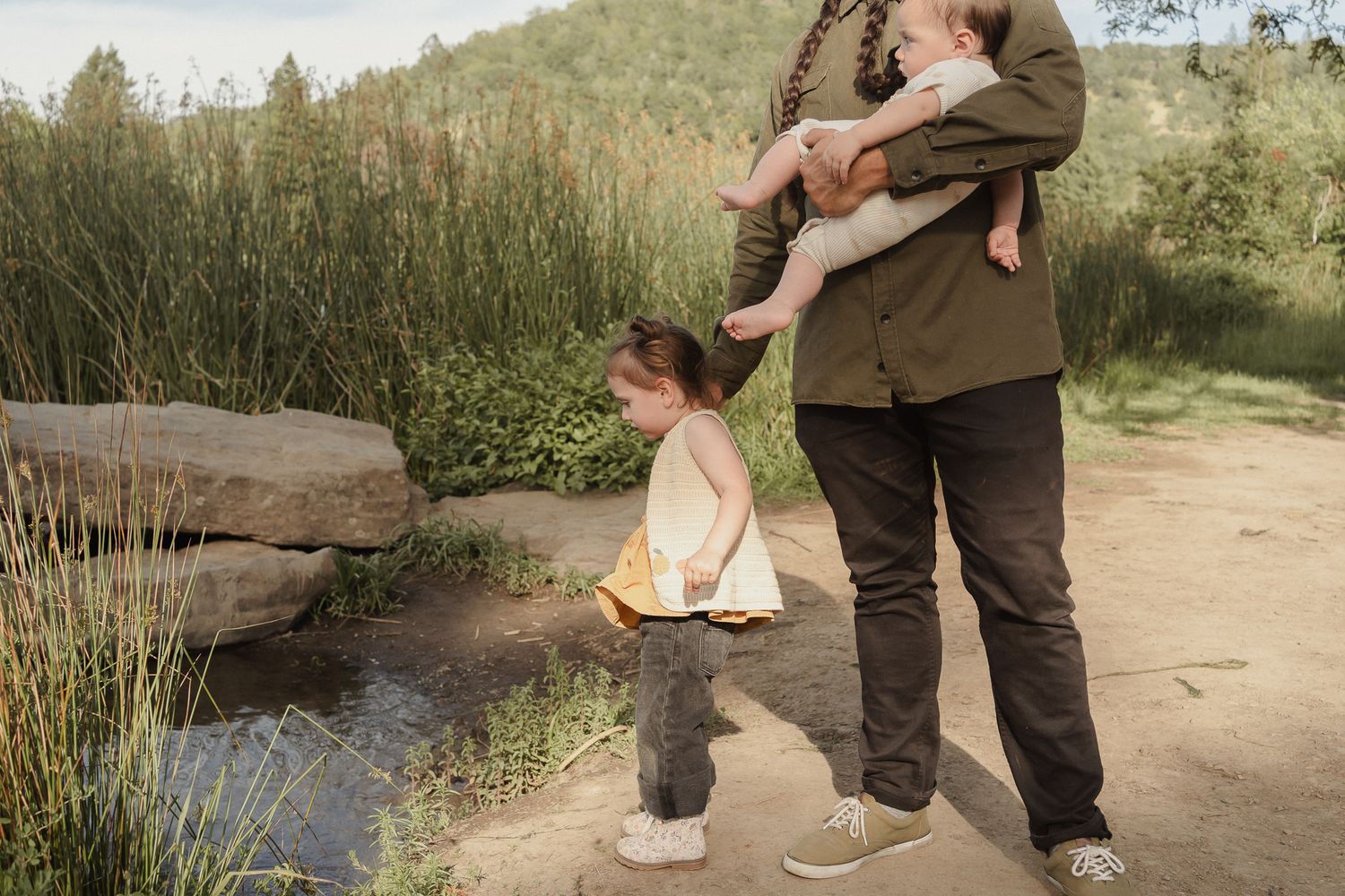 Dad holding a baby standing with a toddler by a pond