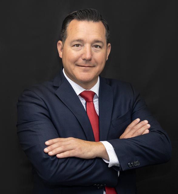 Professional headshot of someone in a navy suit and red tie with arms crossed against dark background.