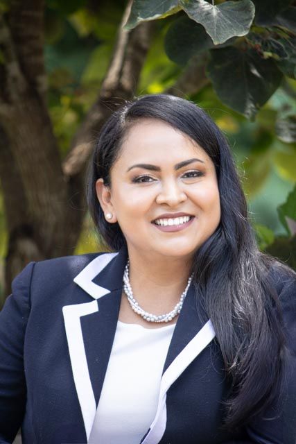 Professional headshot of someone in a navy blazer with white trim and pearl necklace smiling outdoors with foliage background.