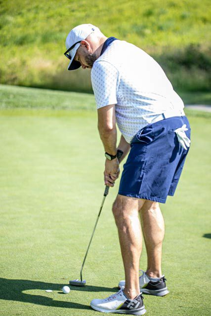 Golfer in white shirt and navy shorts bends down to line up a putt on a sunny green course.