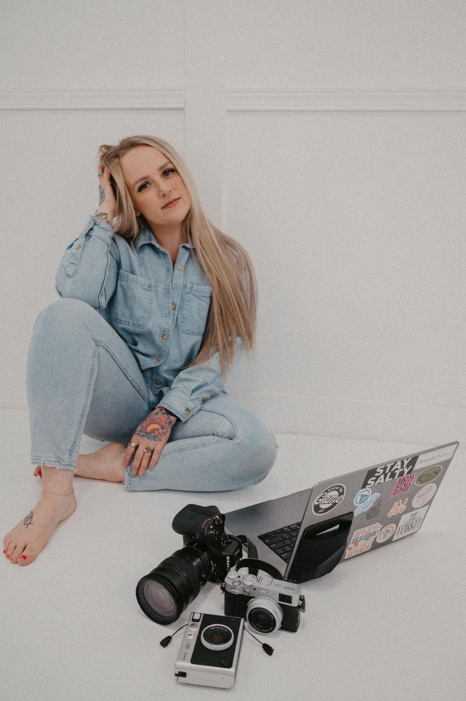 Blonde woman in denim outfit sits on white floor with DSLR cameras and sticker-covered laptop.