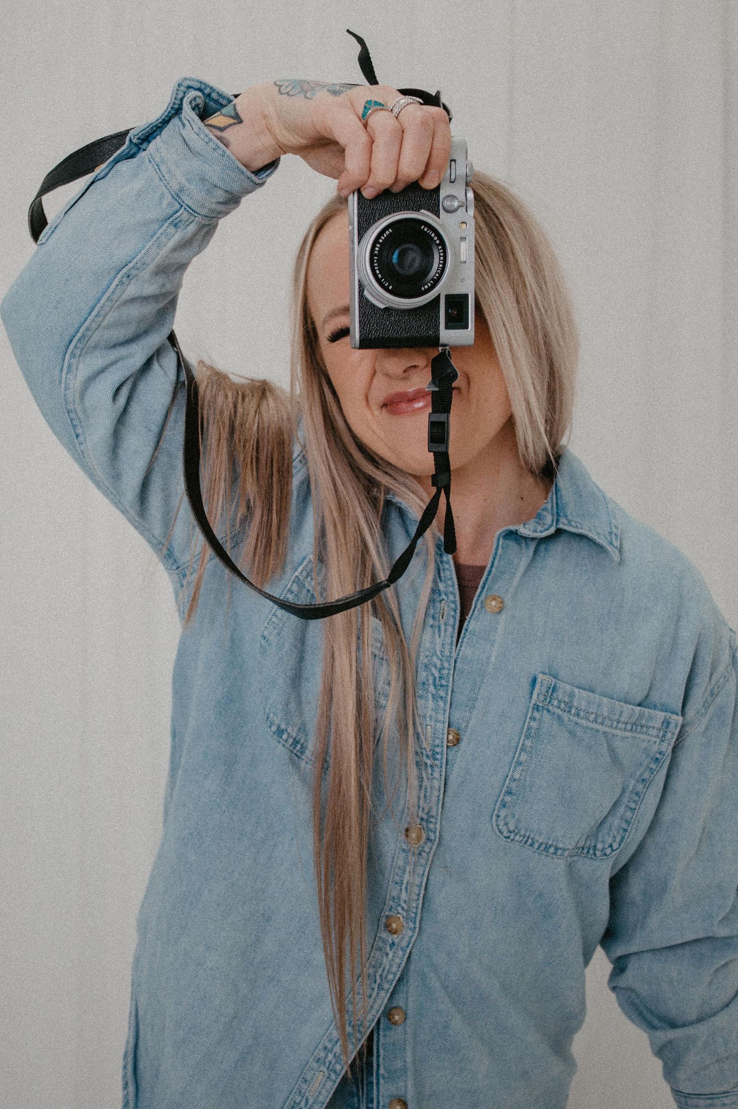 Woman in denim shirt holding a vintage film camera up to her face against a light gray background.