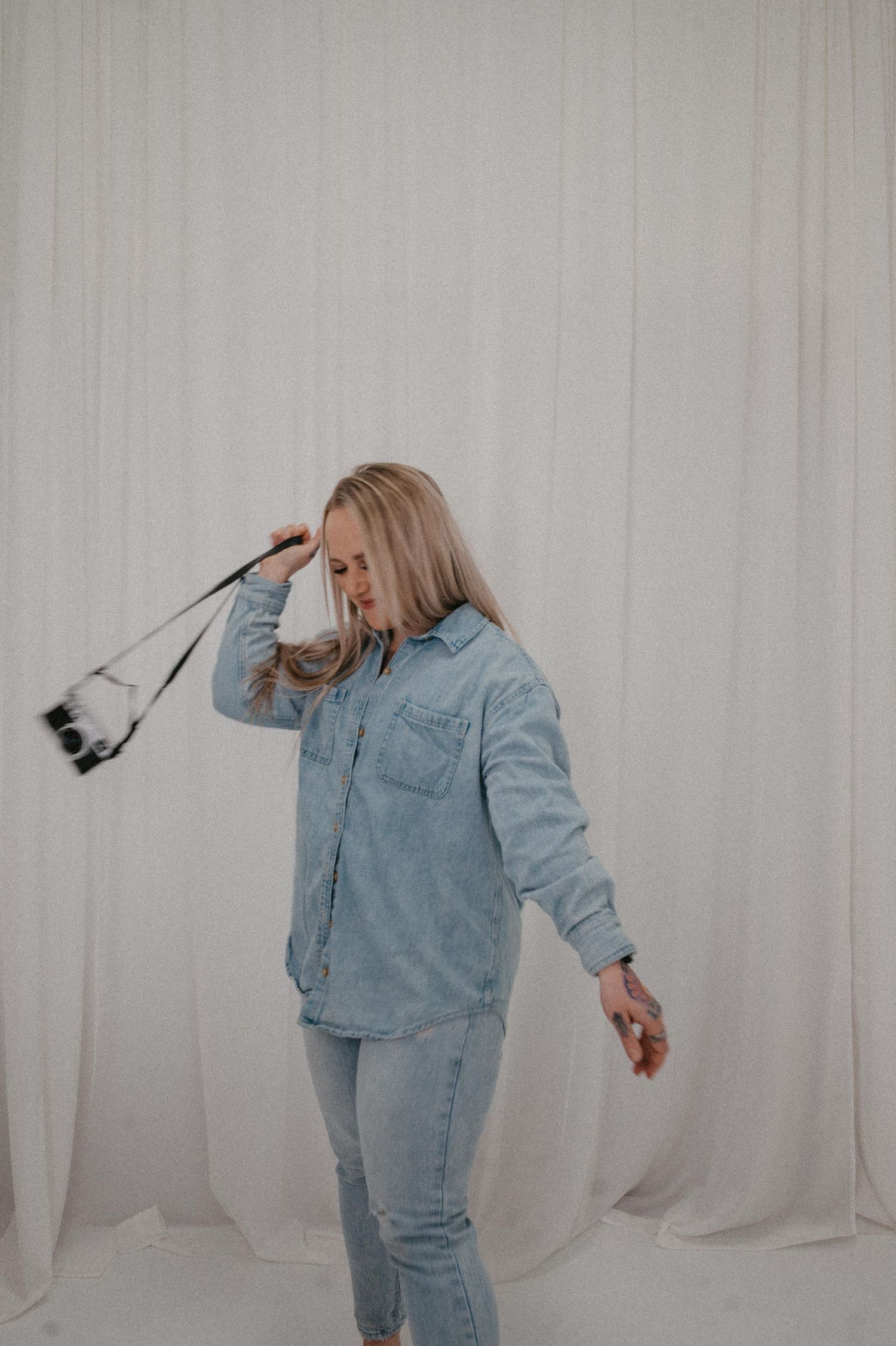 A blonde woman in a denim jacket and jeans holds a camera strap while posing against a white curtain backdrop.