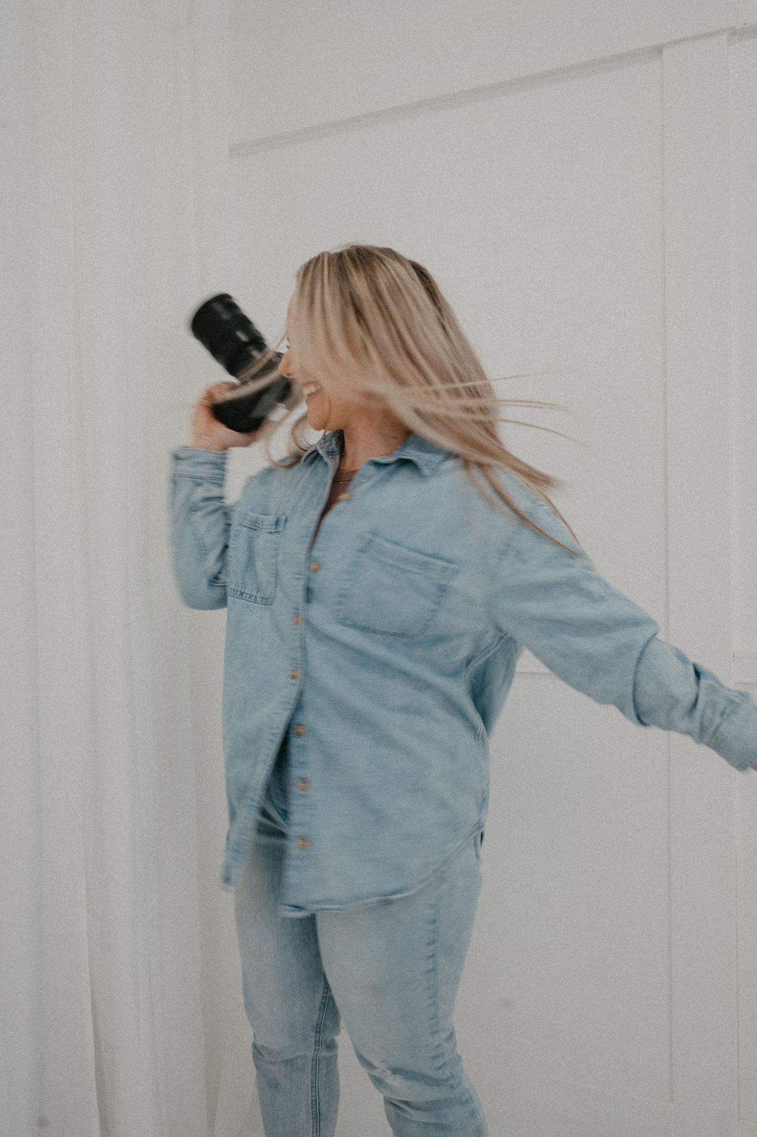 Blonde woman in light blue denim outfit holding a camera up to her face against a white wall.