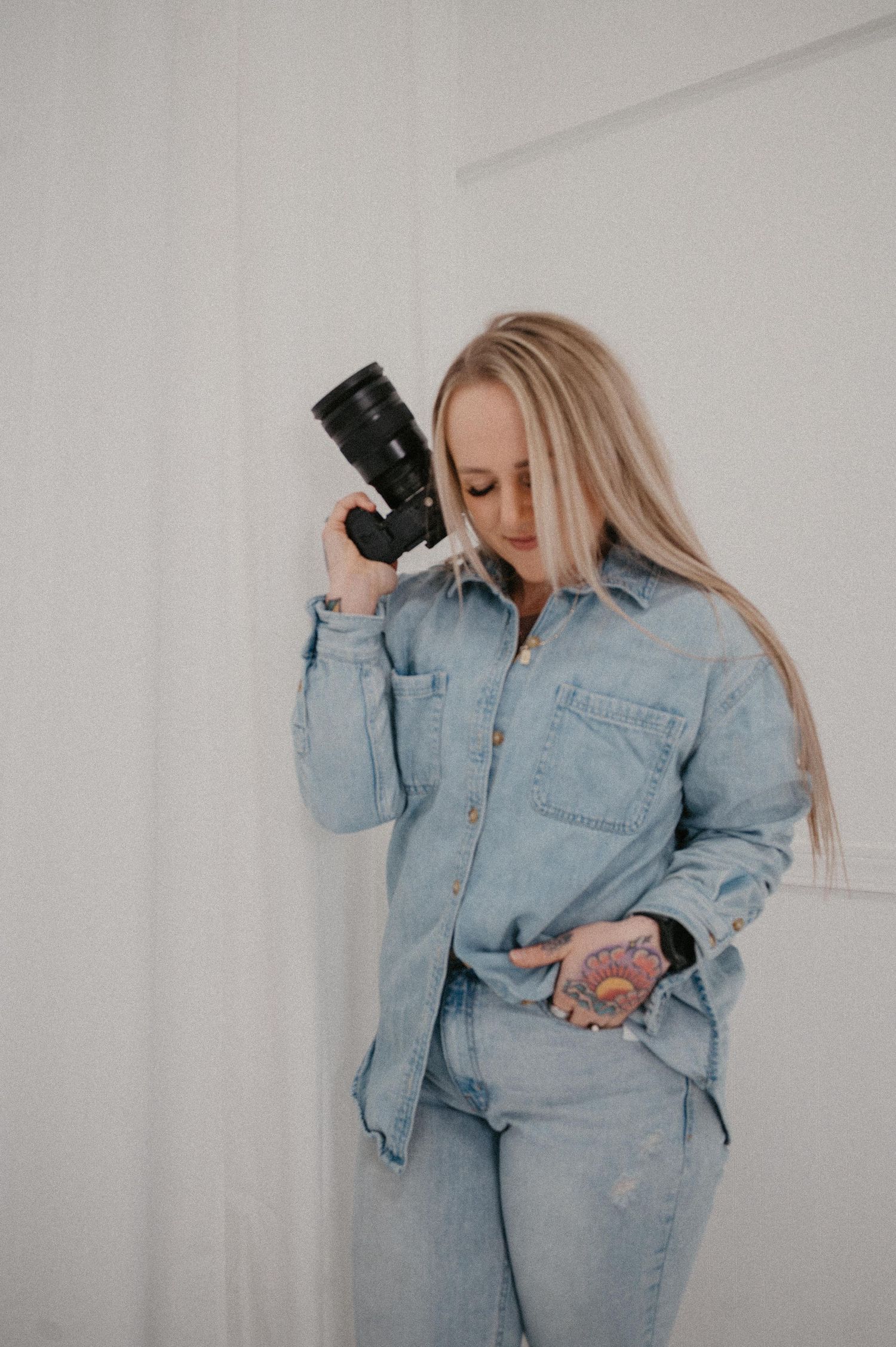 Blonde woman in denim outfit holding a camera against her cheek, posing against a white wall.