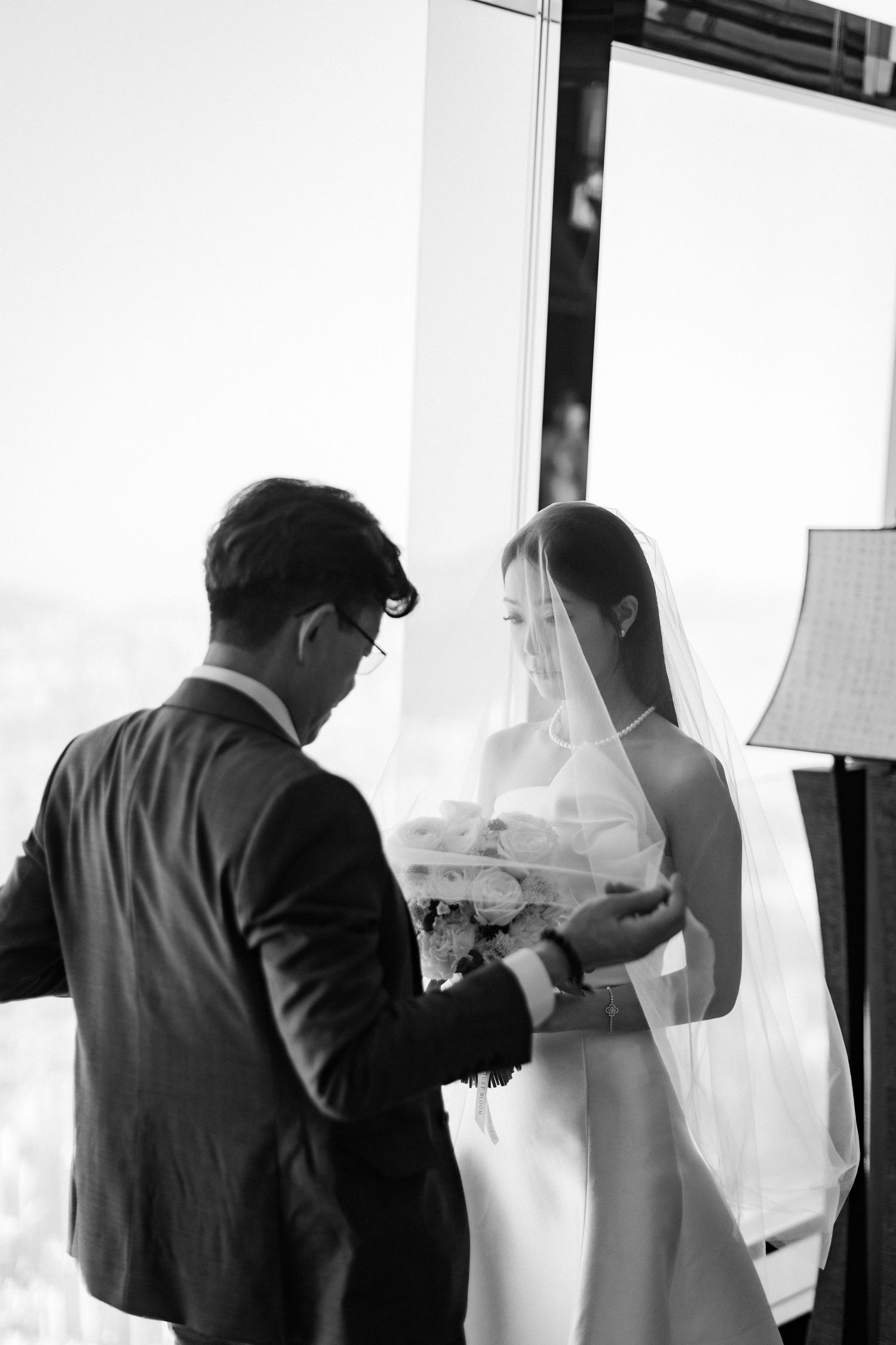 Black and white wedding photo of bride in veil holding bouquet with groom in suit by large window.