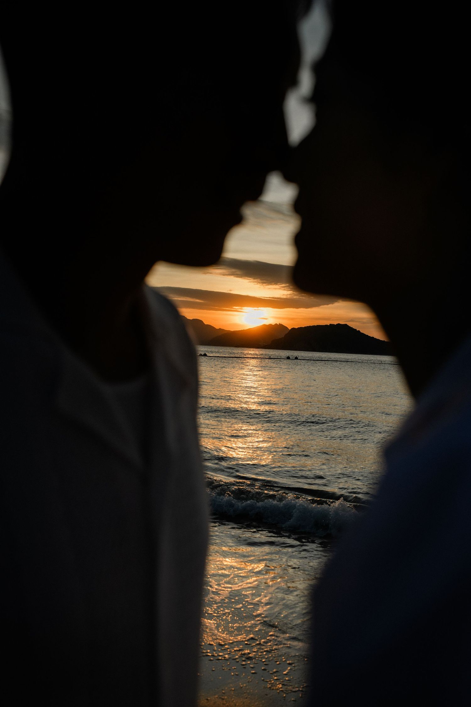 Silhouette of couple facing each other with golden sunset over ocean and mountains behind them.