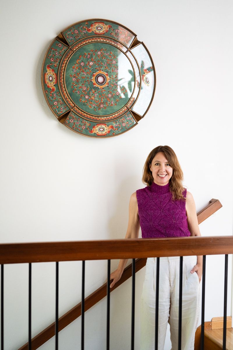 A decorative plate with ornate orange and green patterns hangs above a person in a purple top posing on stairs.