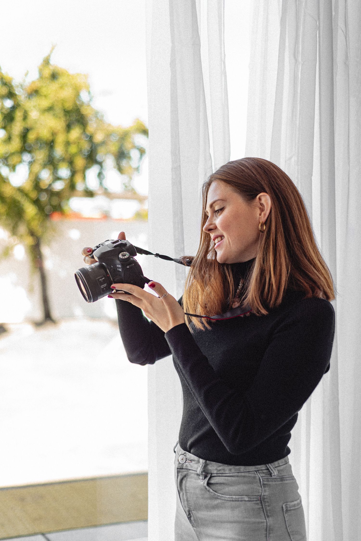 A photographer wearing a black sweater cleans their camera lens while standing next to white curtains.