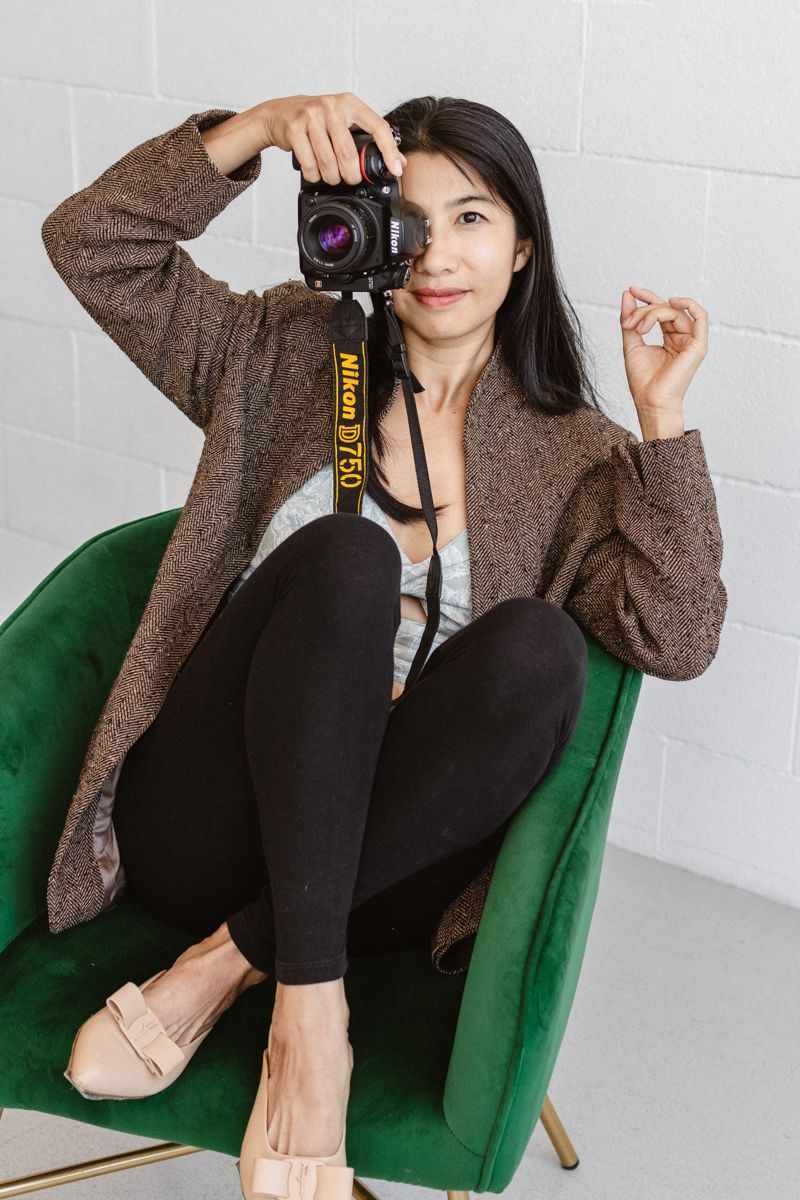Photographer in brown sweater and black pants poses with camera on emerald green velvet chair against white wall.