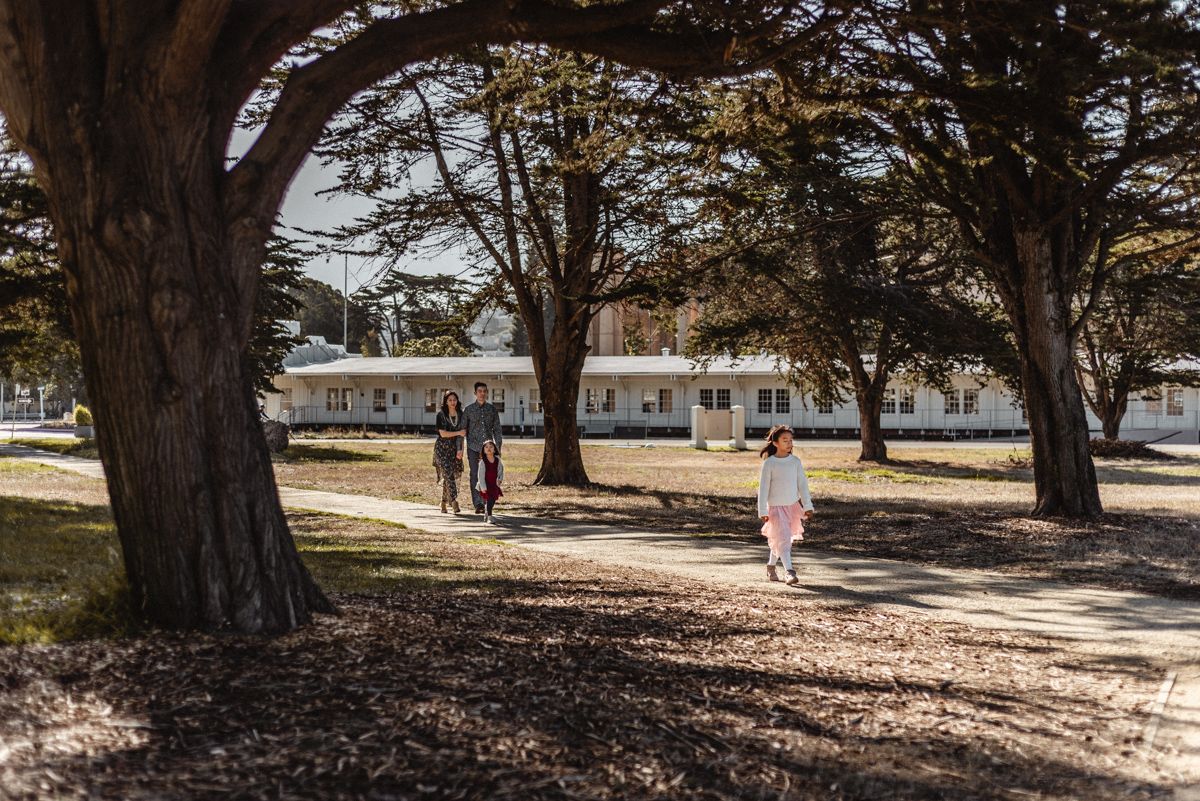 People walking along a tree-lined path near white buildings on a sunny day.