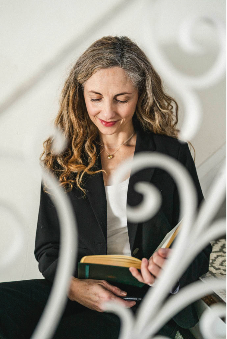 Woman in black blazer sits reading a book in a minimalist white setting with decorative elements.