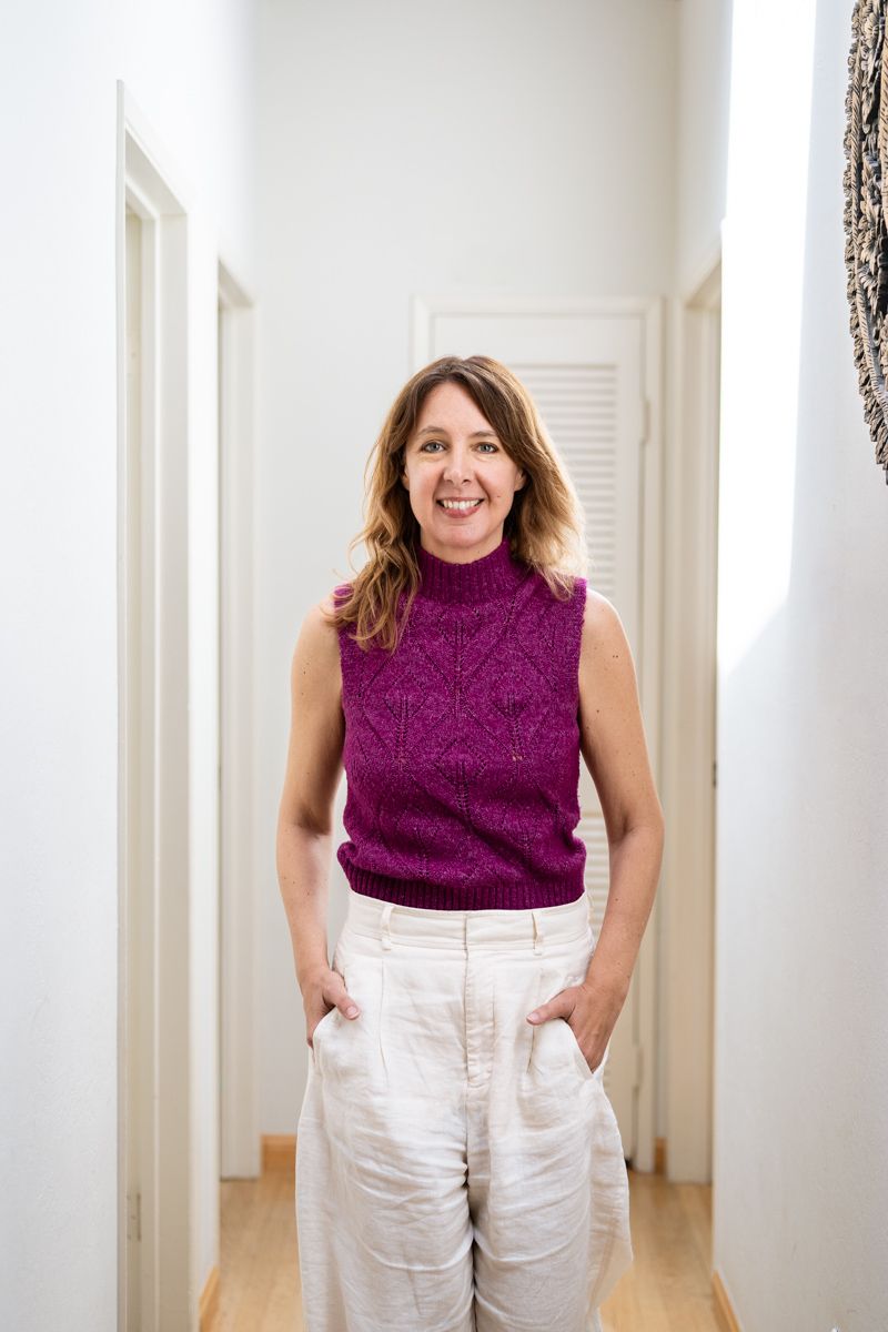 A person wearing a purple sleeveless turtleneck and white pants poses in a bright white hallway.
