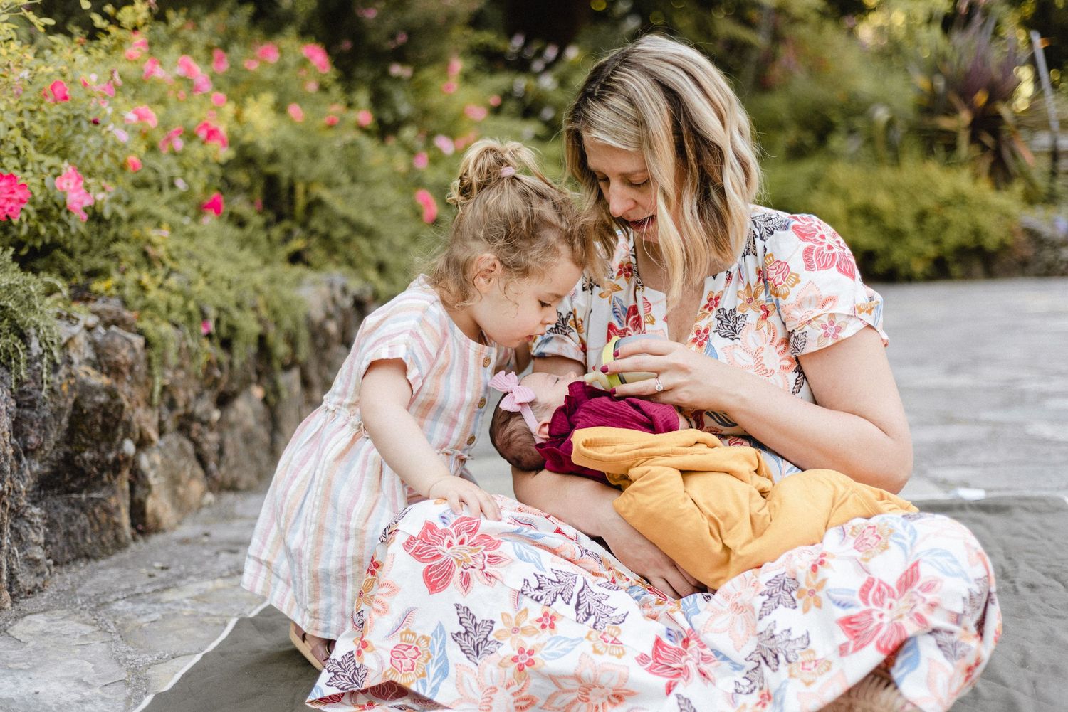 Mother and child sit together on stone wall in garden with blooming flowers.